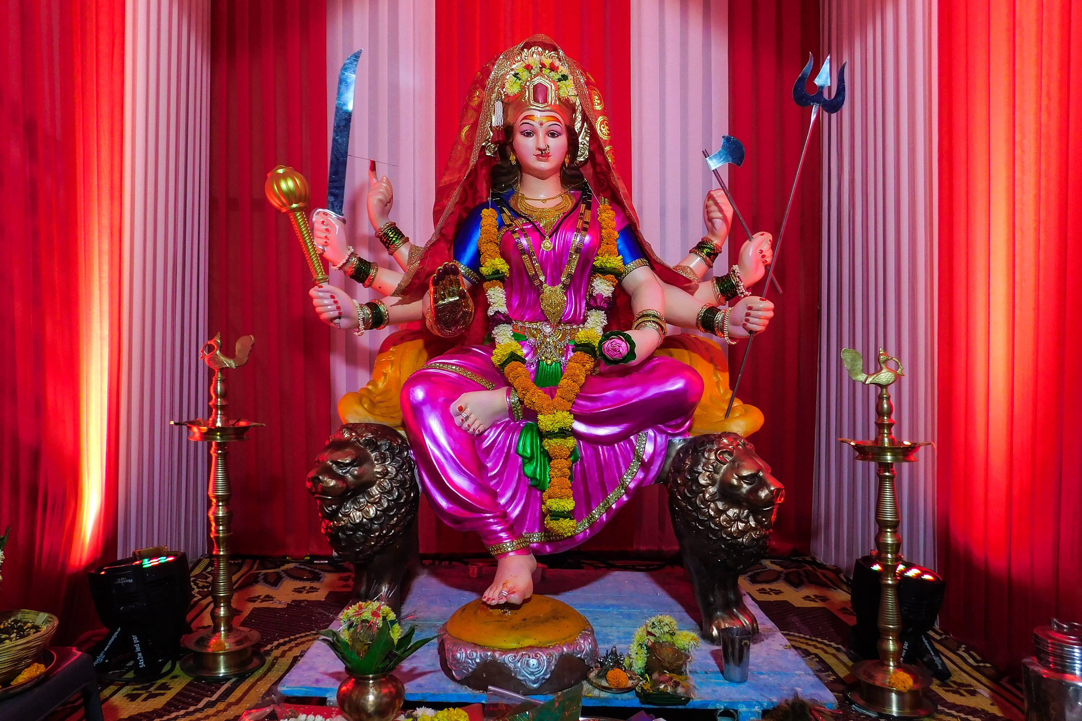 Navratri 桌布 壁紙 — 一座充滿活力的粉紅色多臂印度教女神雕像，飾有黃金首飾和花環，莊嚴地坐在獅子寶座上，背景是紅色窗簾。