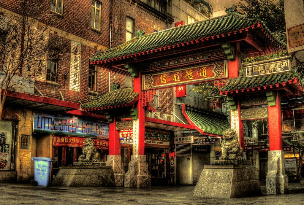 唐人街 桌布 壁紙 — 一座充滿活力的紅綠中國牌坊，配以石獅，矗立於繁華的唐人街街道入口，兩旁是磚砌建築和發光的招牌。