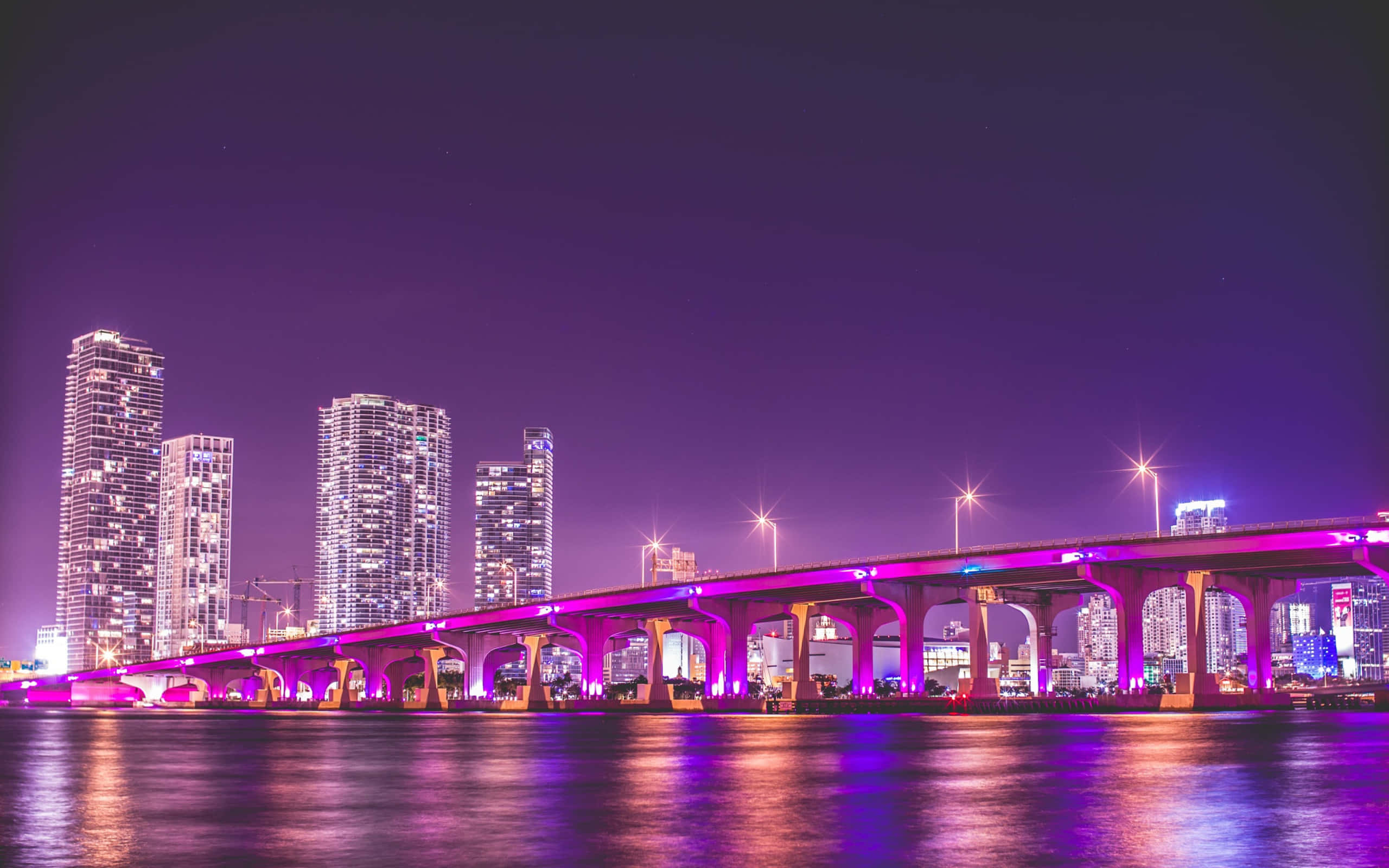 Miami 桌布 壁紙 — 一座充滿活力的紫色橋樑橫跨平靜的水道，背景是深邃的夜空和閃爍的城市摩天大樓。