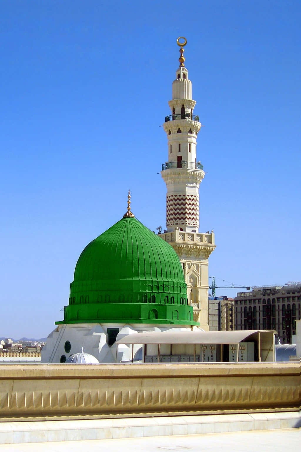 Makkah 桌布 壁紙 — 一座充滿活力的綠色圓頂清真寺，帶有高聳的宣禮塔，矗立在晴朗的藍天下，處於陽光明媚的城市環境中。