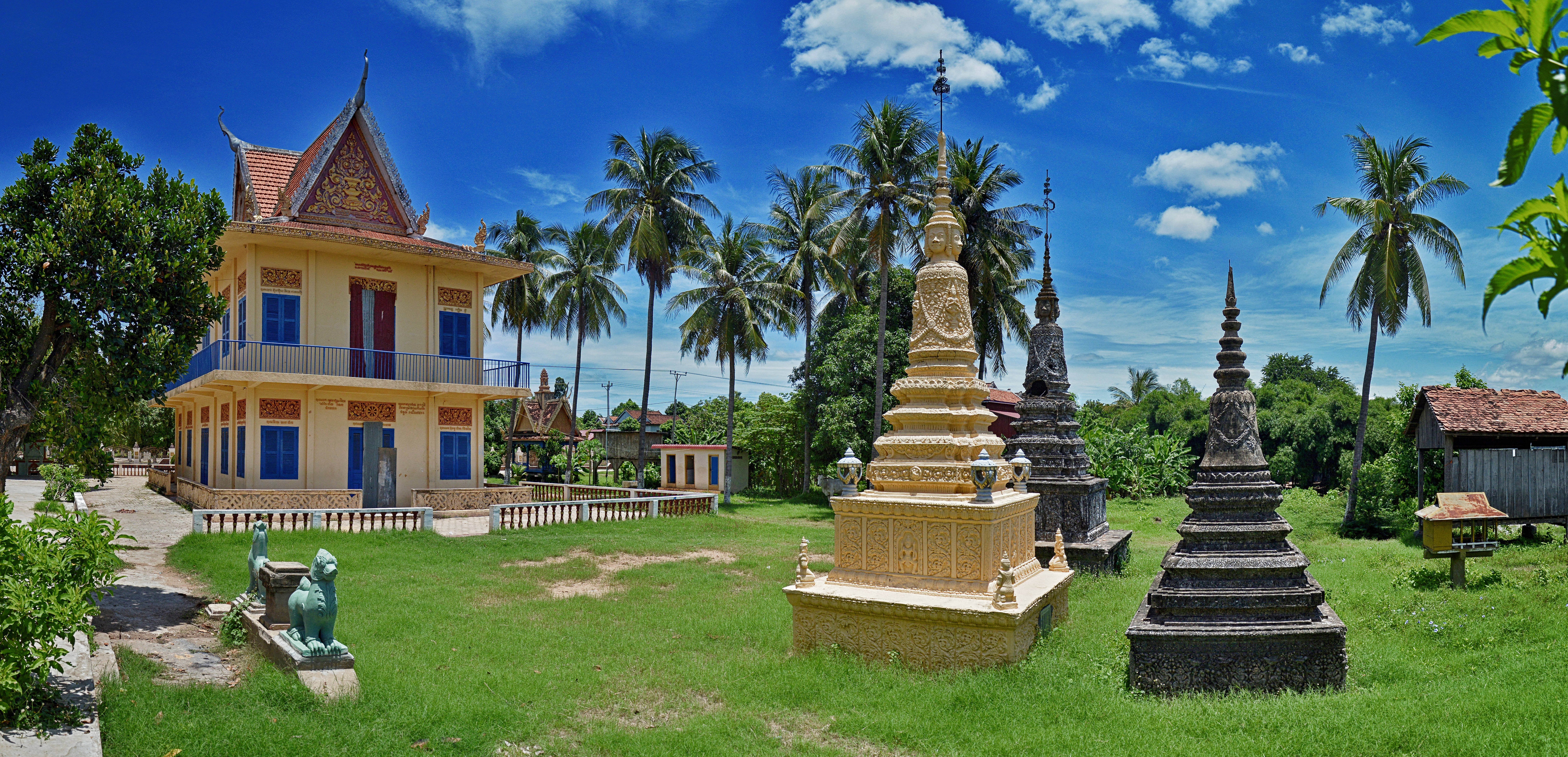 Cambodia 桌布 壁紙 — 一座充滿活力的黃色寺廟建築，帶有藍色百葉窗，矗立在金色和石製佛塔旁，天空湛藍，雲朵稀疏。