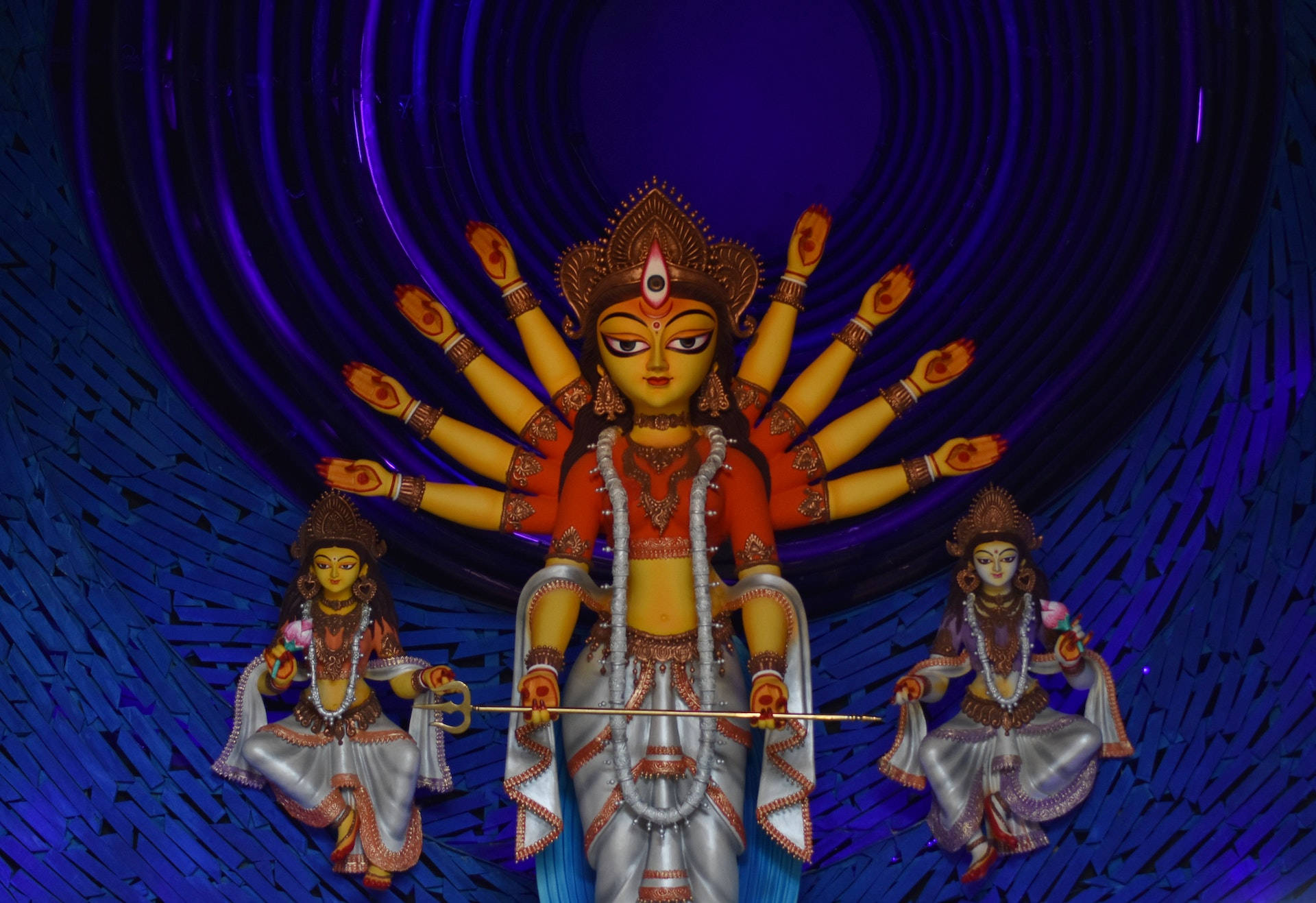 Durga 桌布 壁紙 — 一座光芒四射的多臂女神雕像與兩位侍者，矗立在旋轉的深藍色螺旋背景前。