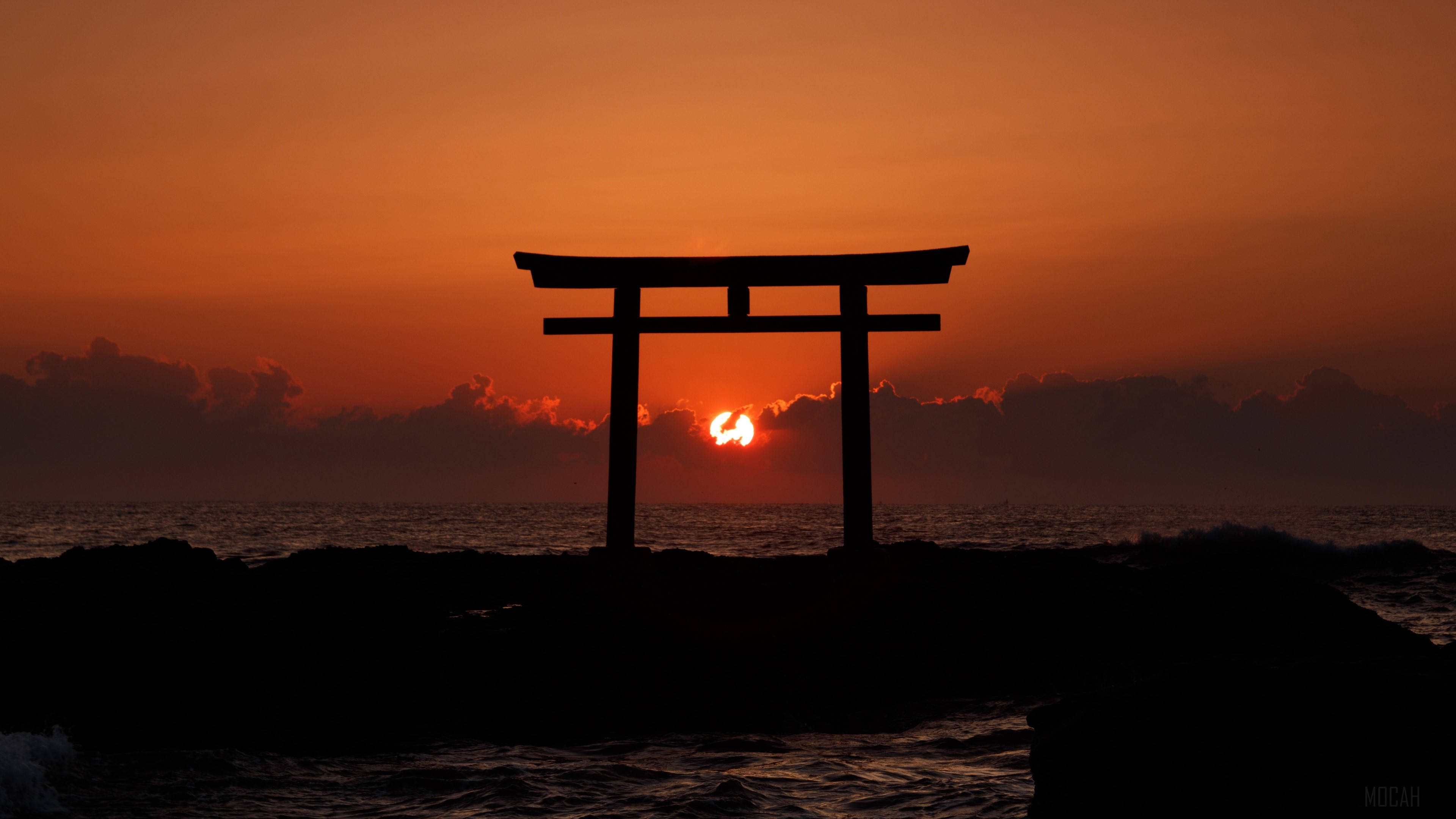 Torii 桌布 壁紙 — 一座剪影風格的日本鳥居矗立在岩石海岸上，夕陽在其後方以火橙色沉入海洋。