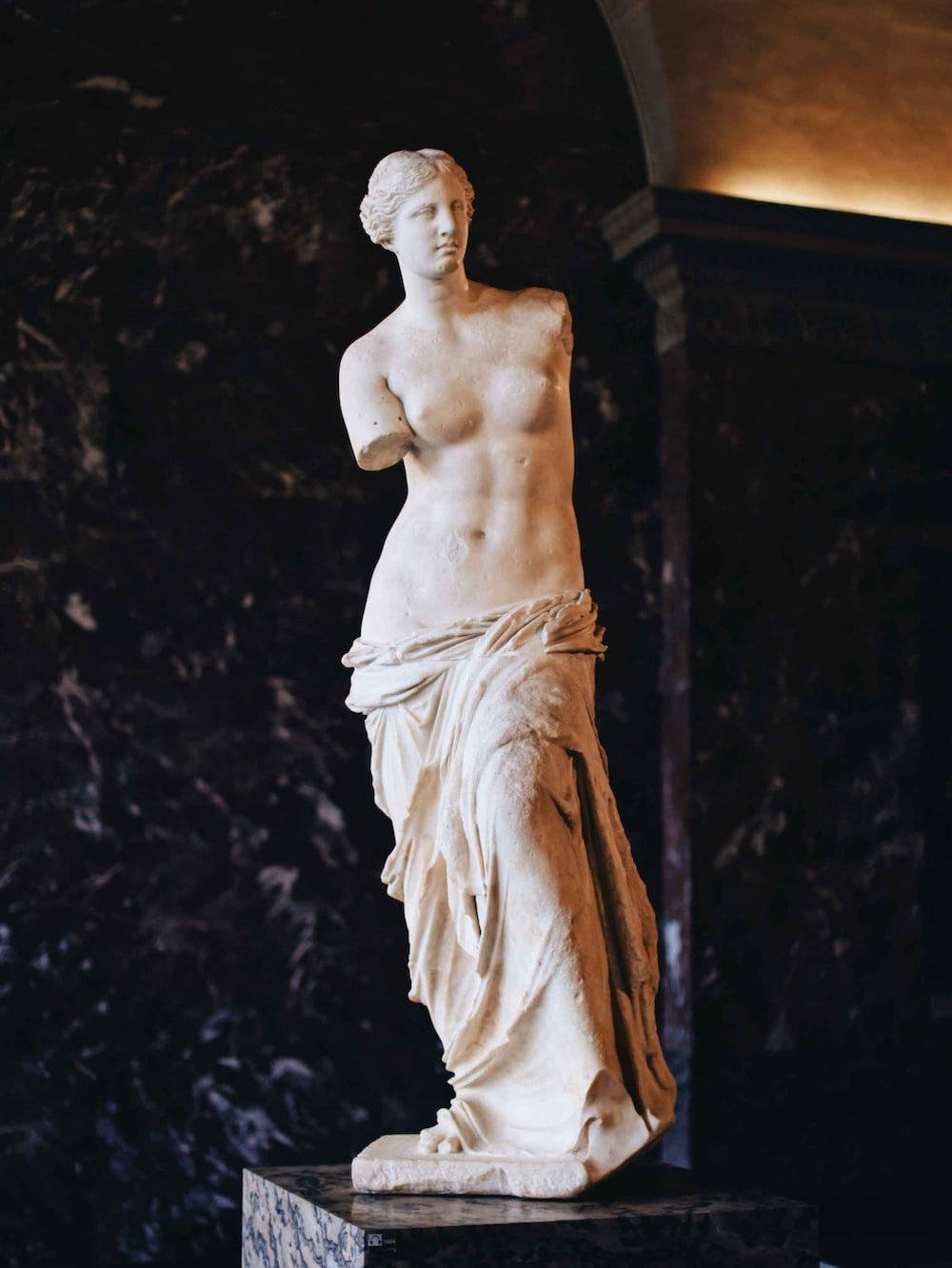 Statue Aesthetic 桌布 壁紙 — 一座古典的米洛的維納斯大理石雕像優雅地立於基座上，背景是深色帶紋理的大理石牆，在溫暖的環境燈光下。