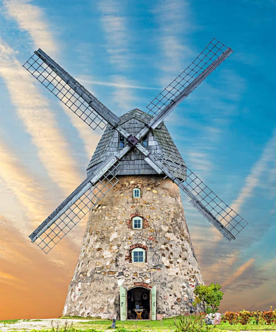 windmill 桌布 壁紙 — 一座古樸的石磨風車高聳在色彩斑斕的日落天空下，天空有條紋狀的雲朵，下方是綠草如茵。