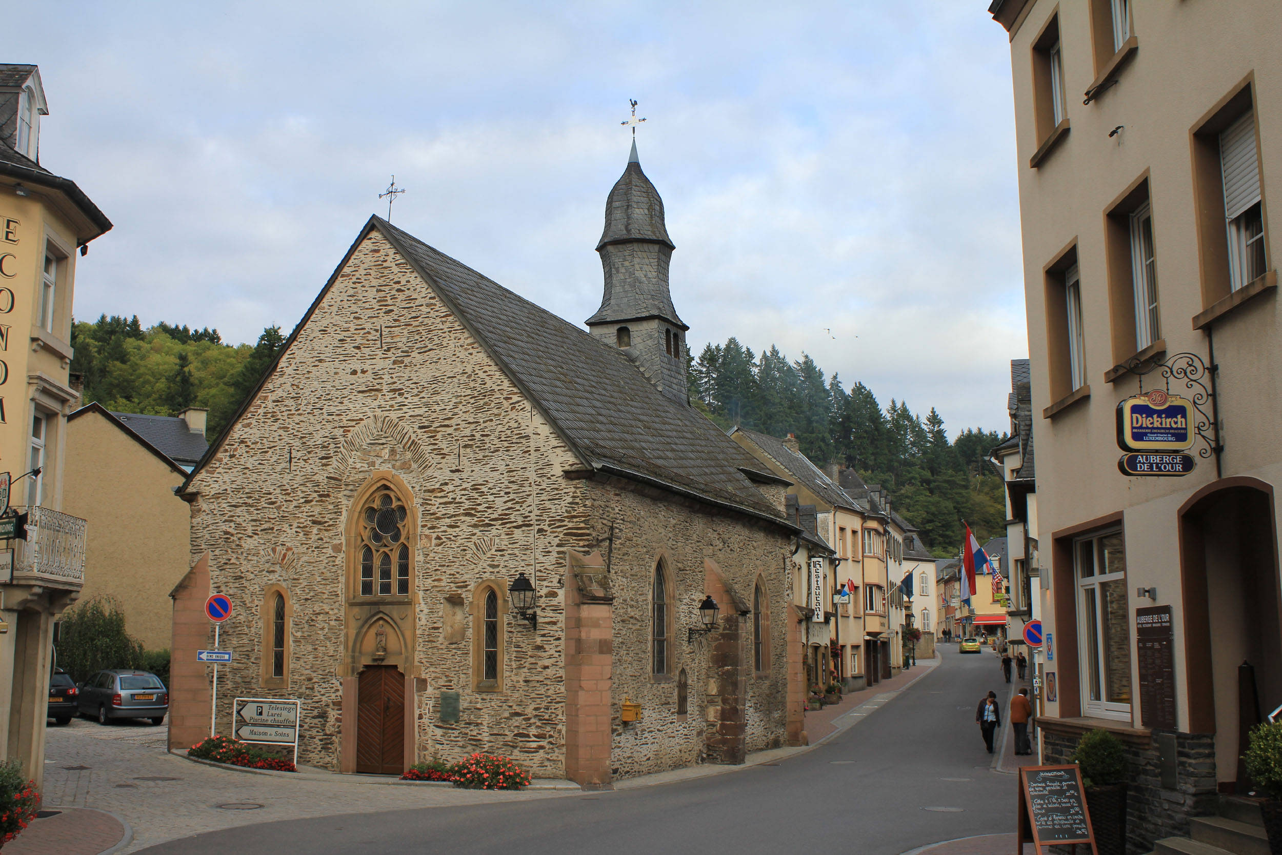Luxembourg 桌布 壁紙 — 一座古雅的石砌教堂，擁有石板屋頂和鐘樓，矗立在歐洲小鎮的街角，兩旁是建築物和樹木成蔭的山丘，天空多雲。