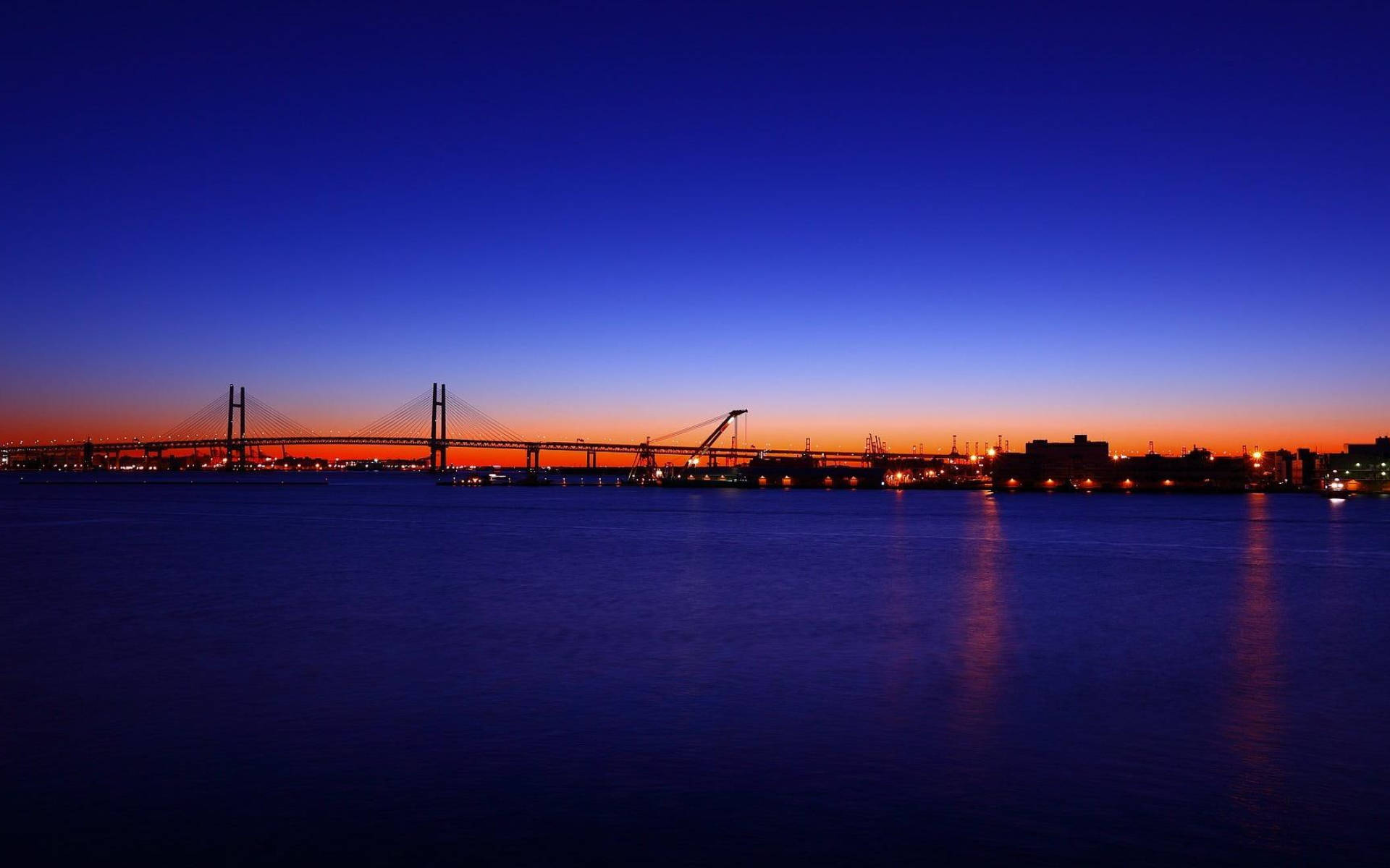 Yokohama 桌布 壁紙 — 一座吊橋在平靜的水面上，以深藍色和火橙色漸變的天空為背景，呈現出令人驚嘆的黃昏景色。