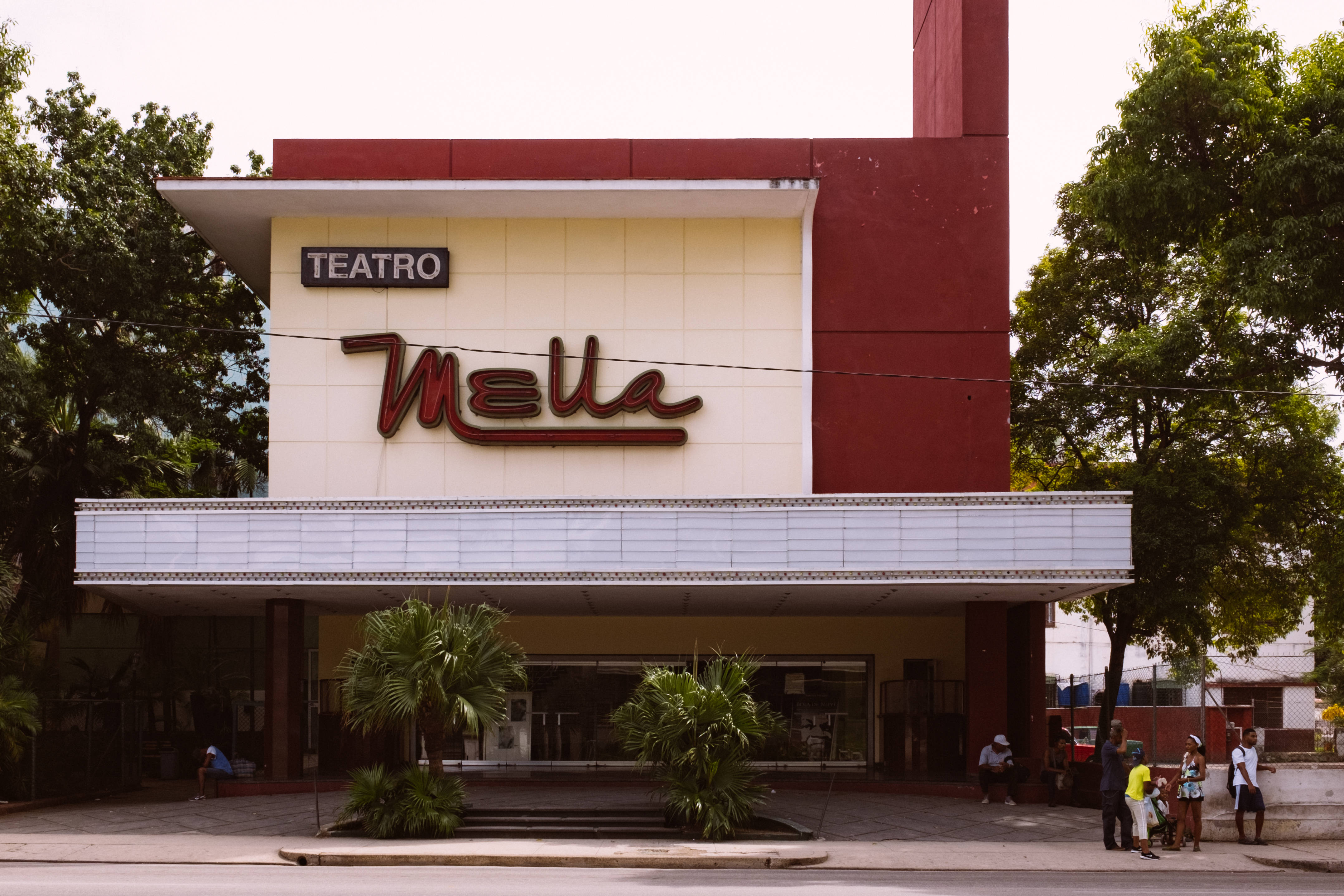 Cuba 桌布 壁紙 — 一座名為Teatro Mella的復古紅色和奶油色劇院，在晴朗的天空下，入口附近有棕櫚樹和人。