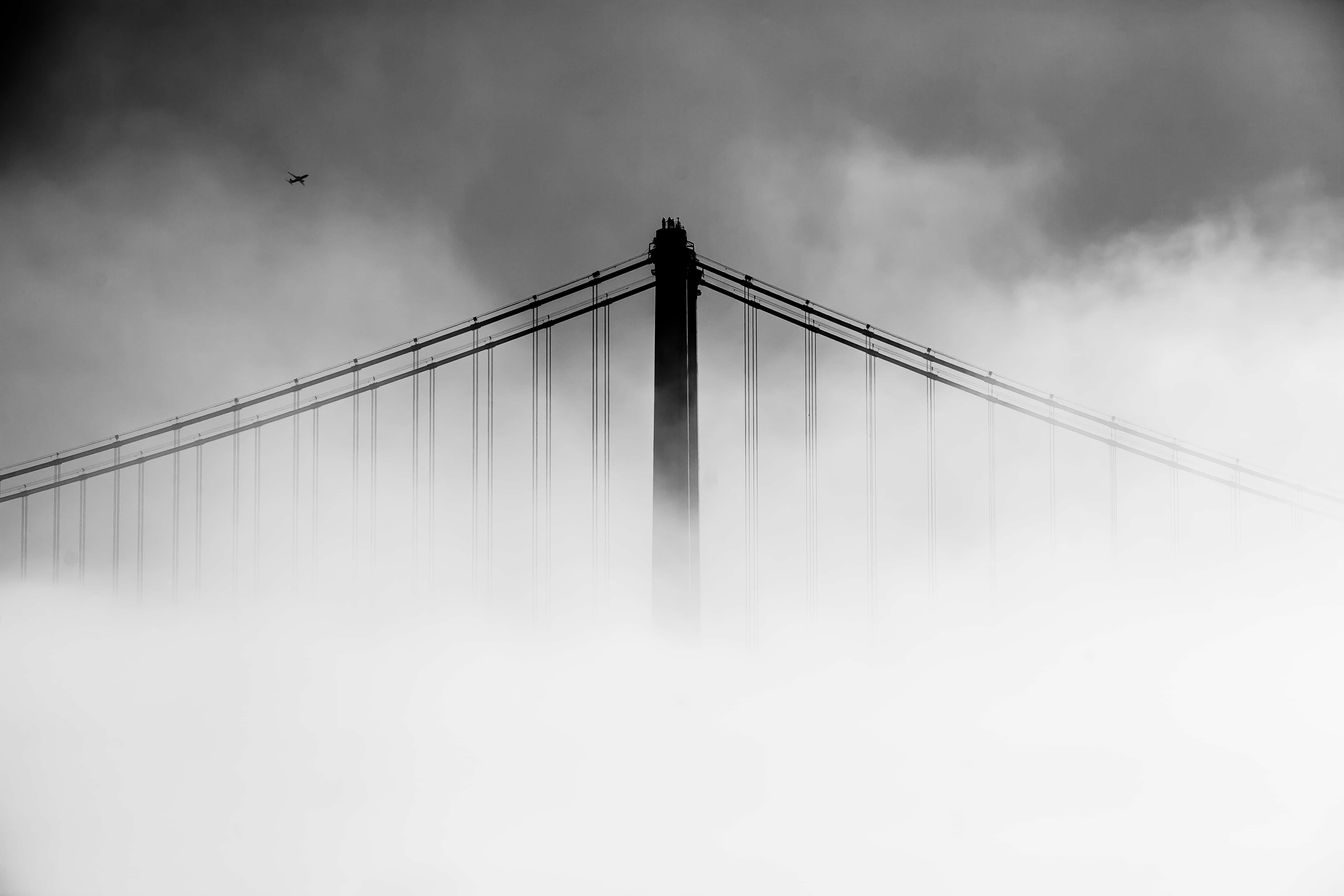 San Francisco Black And White 桌布 壁紙 — 一座單色吊橋穿透濃霧，一架孤獨的飛機在陰雲密佈的天空上方飛過。