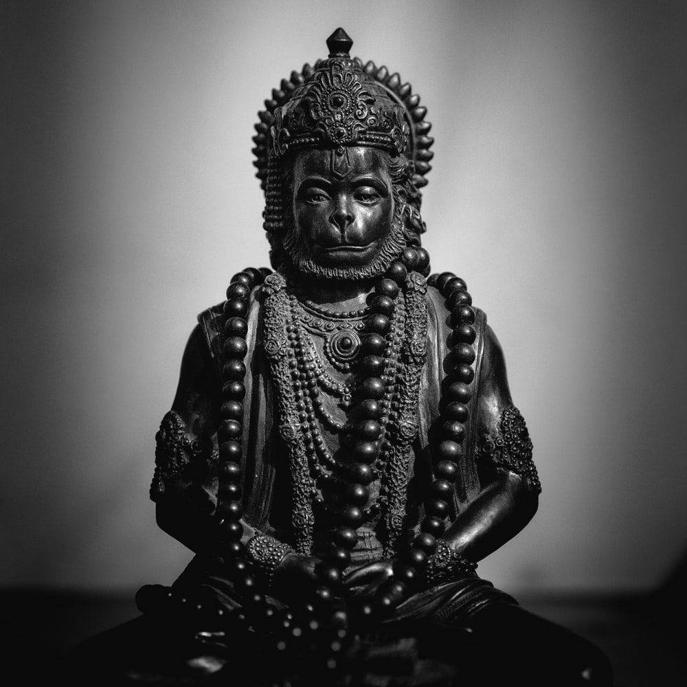 hanuman 4K HD 桌布 壁紙 — 一座單色調的寧靜猴神雕像，飾有複雜的珠寶和珠子，以冥想姿勢坐在柔和的漸變背景前。