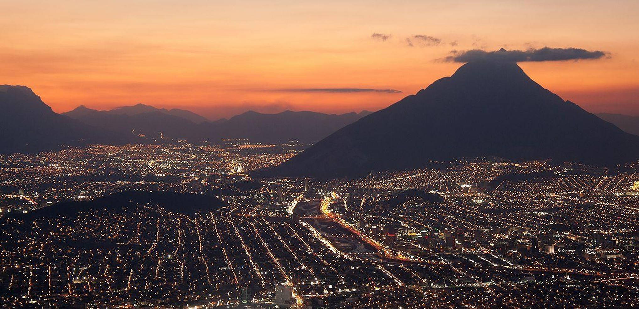 Monterrey 桌布 壁紙 — 一座在黃昏時分蔓延的城市，發光的街燈和黑暗的山脈輪廓映襯著橙色的天空。