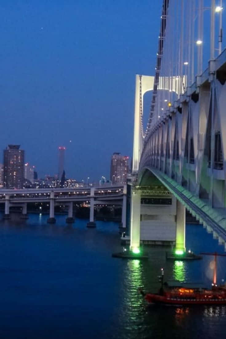 Rainbow Bridge 桌布 壁紙 — 一座在黃昏時發光的橋樑倒映在平靜的水面上，一艘亮燈的船隻從橋下穿過，背景是城市天際線。