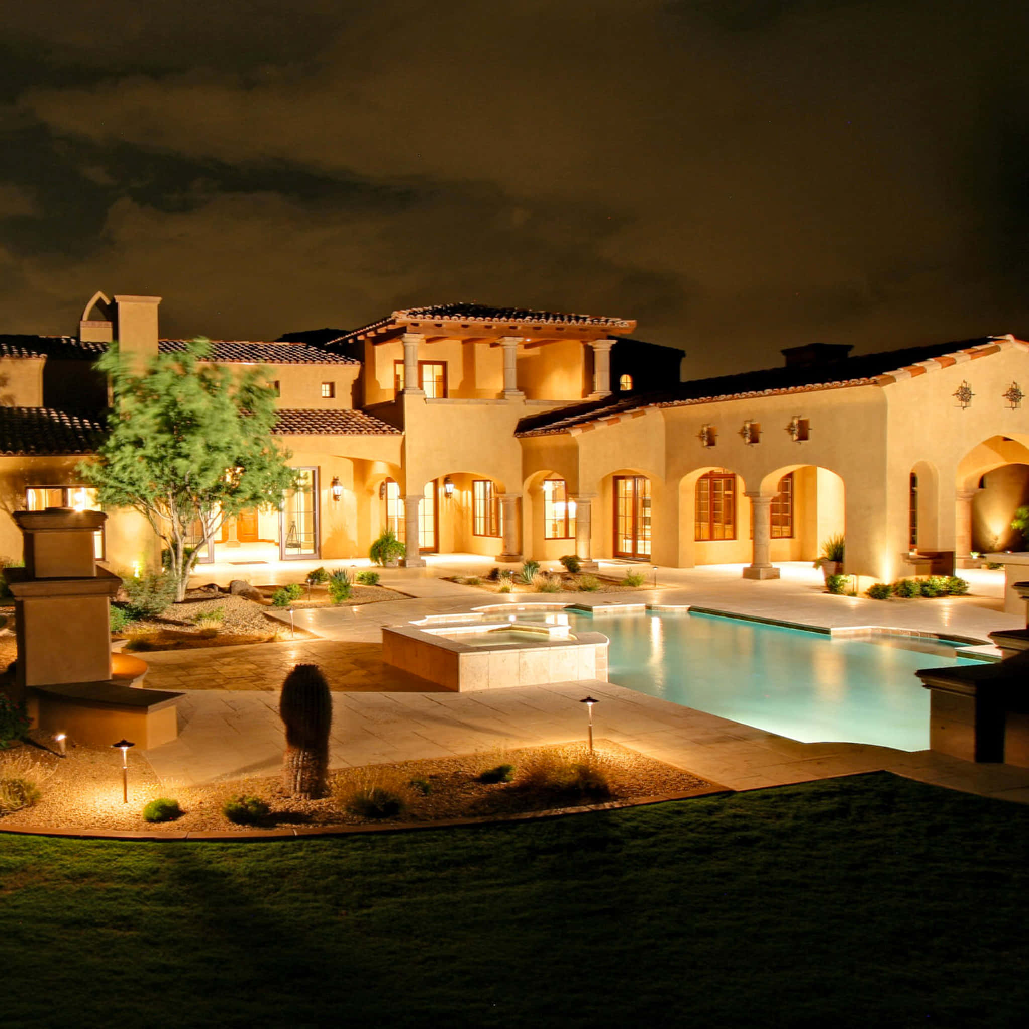 Mansion 桌布 壁紙 — 一座地中海風格的大宅在夜晚溫暖地發光，庭院泳池在黑暗的天空下反射著周圍的燈光。