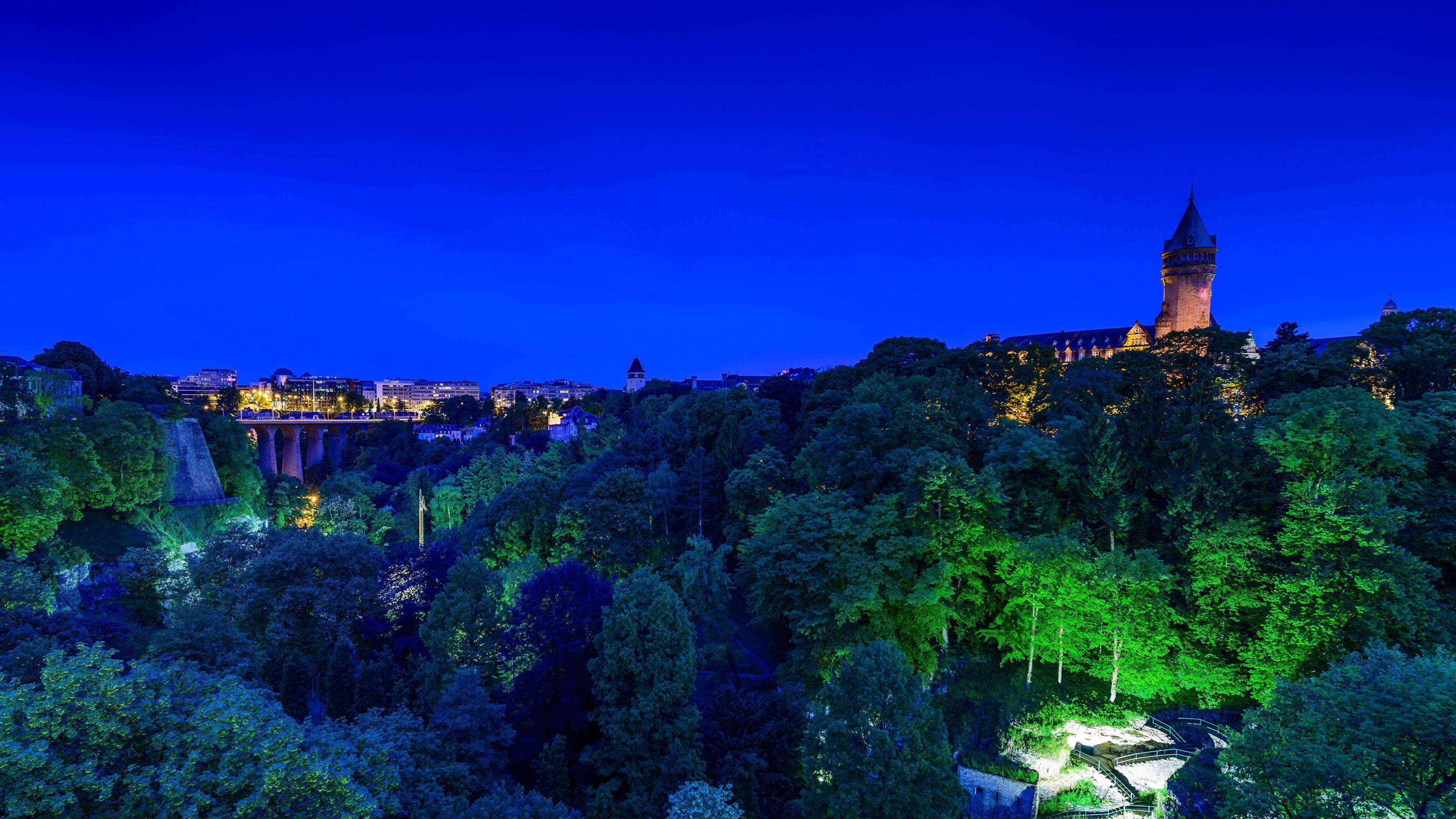 Luxembourg 桌布 壁紙 — 一座城堡坐落在樹木繁茂的山丘上，在深藍色的暮色天空中溫暖地發光，而城市的燈光在下方閃爍。