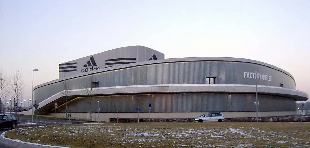 愛迪達 桌布 壁紙 — 一座大型弧形 adidas 工廠直銷建築，帶有金屬壁板和前面有雪塵的草地，在黃昏時分。
