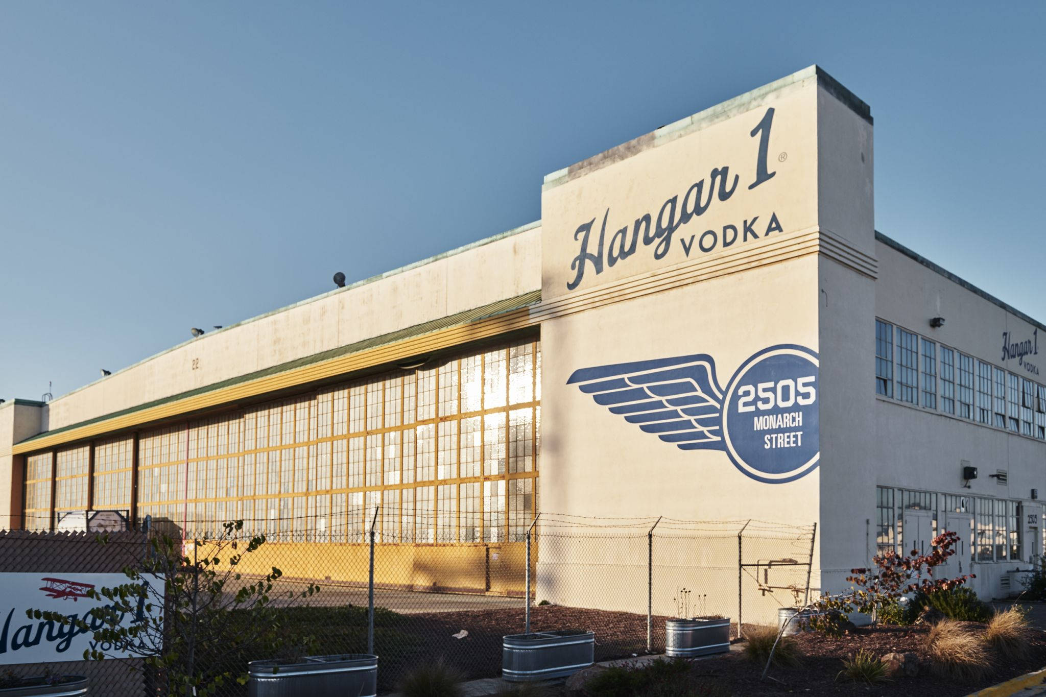 alcohol 桌布 壁紙 — 一座大型米色工業建築，帶有 'Hangar 1 Vodka' 品牌和翅膀標誌，在晴朗的藍天下。