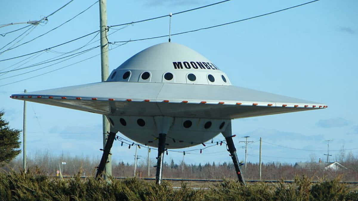 外星人 桌布 壁紙 — 一座大型銀色UFO形狀的建築物，上面標有「MOONBEAM」，矗立在戶外，頭頂有電線，天空清澈湛藍。