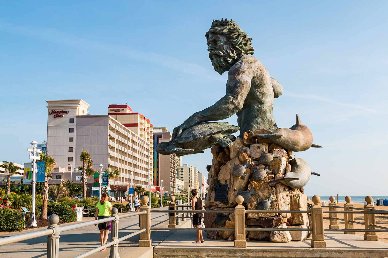 Statue Aesthetic 桌布 壁紙 — 一座大型青銅鬍鬚海神雕像，手持海豚，立於岩石基座上，旁邊是木板路，有飯店和棕櫚樹，天空清澈湛藍。