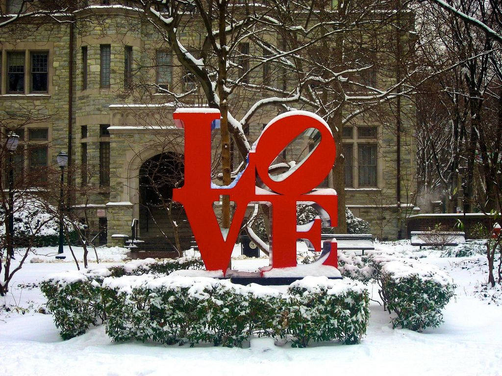 Building 桌布 壁紙 — 一座大膽的紅色LOVE雕塑矗立在冬季校園庭院中，被雪覆蓋，周圍有石頭建築和光禿禿的樹木。