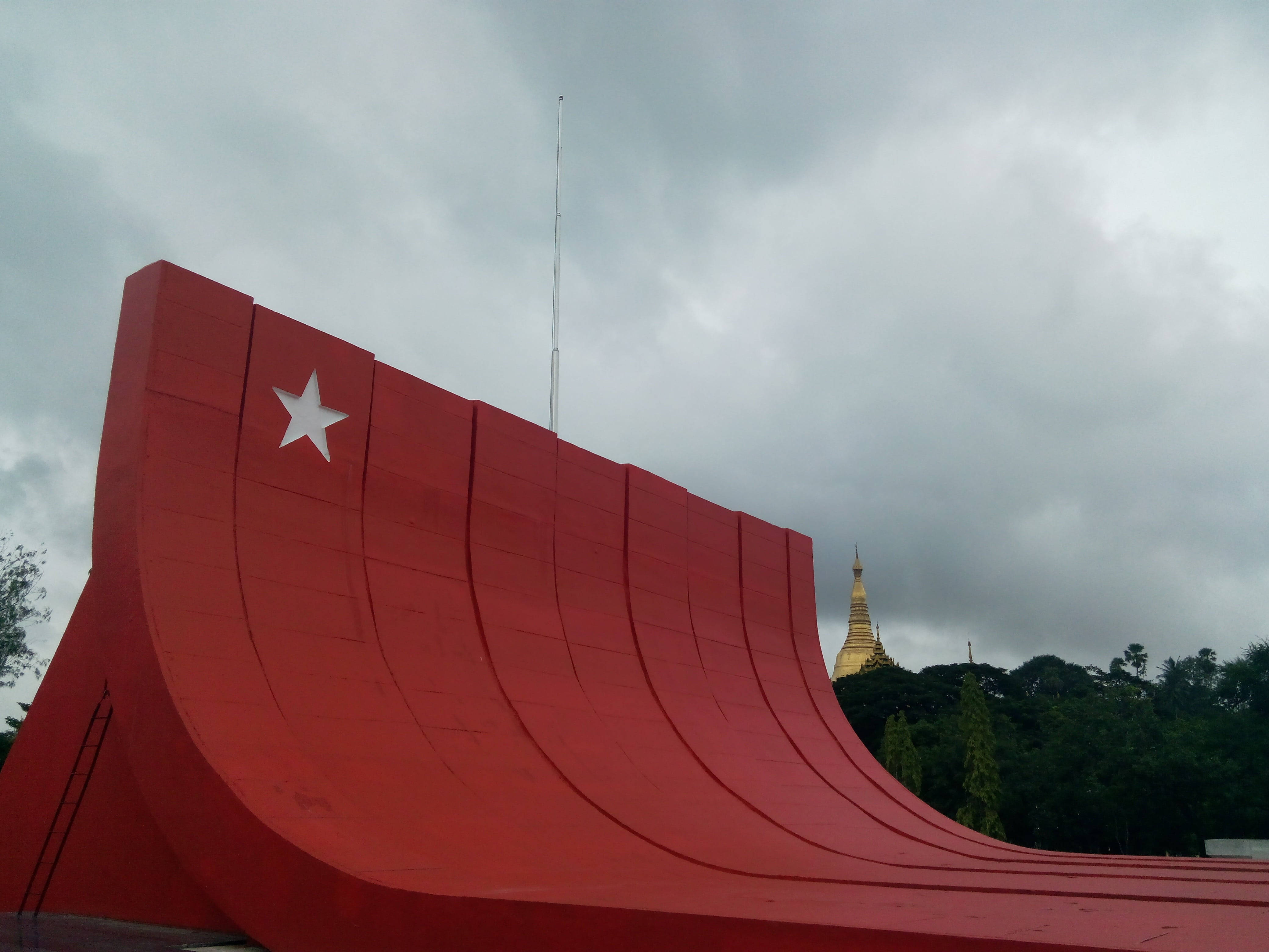 Yangon 桌布 壁紙 — 一座大膽的紅色弧形紀念碑，帶有白色星星，矗立在陰天之下，遠處的金色寶塔在樹叢中若隱若現。