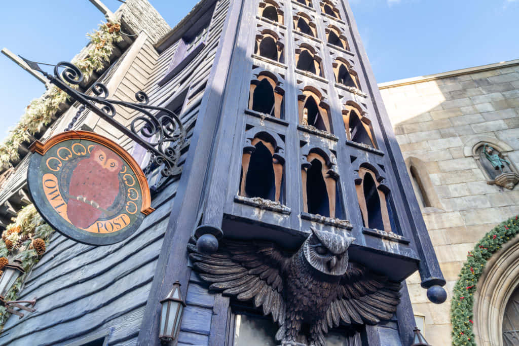 The Hogwarts Owlery 桌布 壁紙 — 一座奇幻的深藍色貓頭鷹主題郵局建築，帶有華麗的貓頭鷹雕刻和節日花環，在清澈的藍天下。