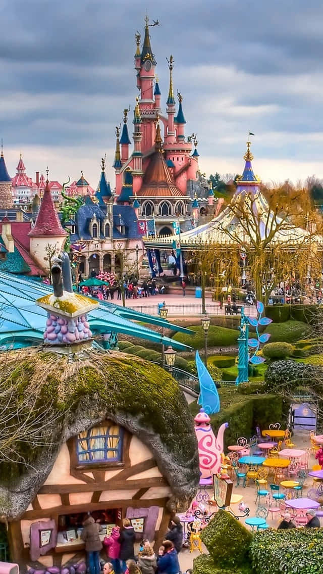 Disneyland Castle 桌布 壁紙 — 一座奇幻的童話城堡，擁有色彩繽紛的塔樓和藍色屋頂，聳立在充滿遊客和粉彩茶館桌子的主題公園之上。