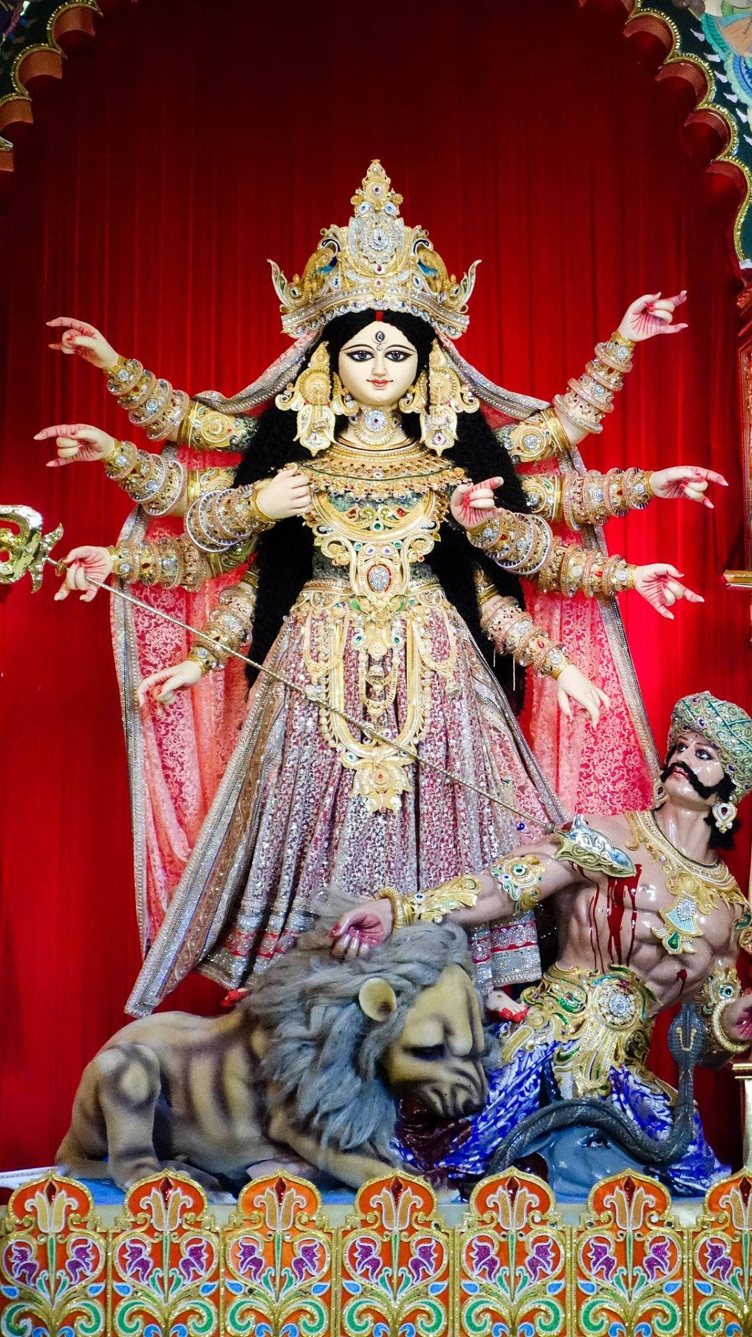 durga mata HD 桌布 壁紙 — 一座威嚴的十臂金色女神像，在華麗的紅色帷幕背景下，勝利地立於被擊敗的惡魔和獅子之上。