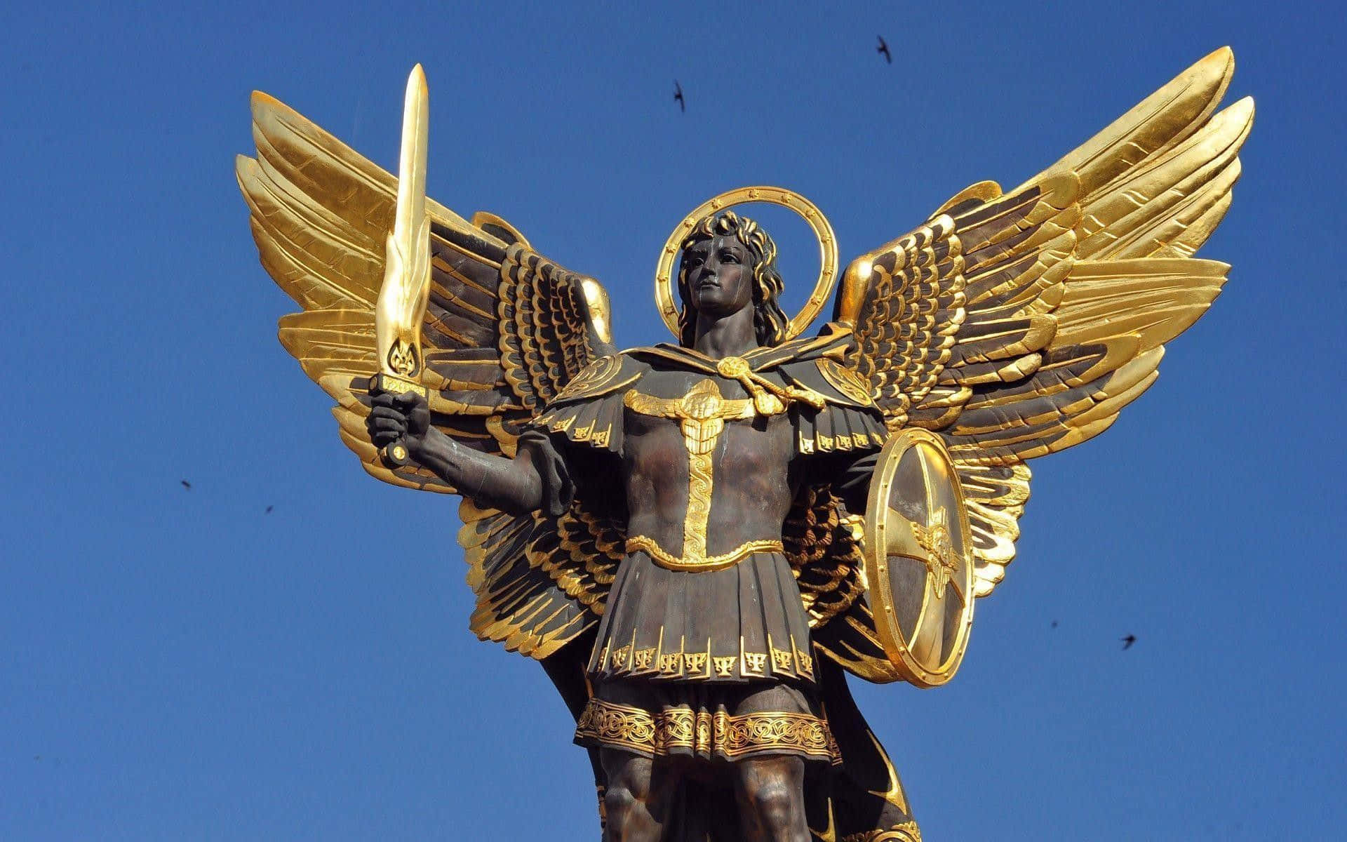 Saint Michael 桌布 壁紙 — 一座威嚴的金翼天使雕像身穿盔甲，手持劍和盾牌，背景是晴朗的藍天，有鳥兒飛翔。