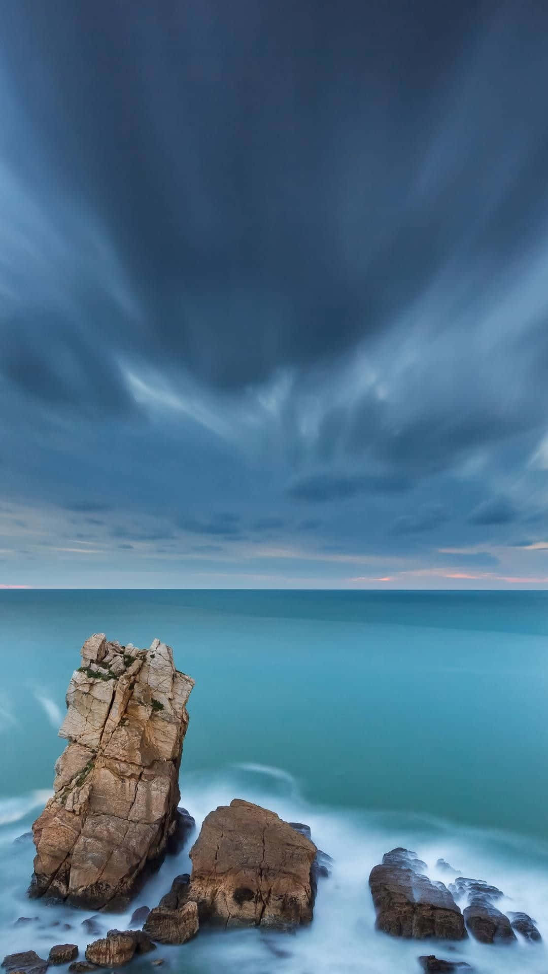 iPhone Home Screen 桌布 壁紙 — 一座孤獨崎嶇的岩石聳立在旋轉的藍天下，光滑的藍綠色水流繞過它的底部。