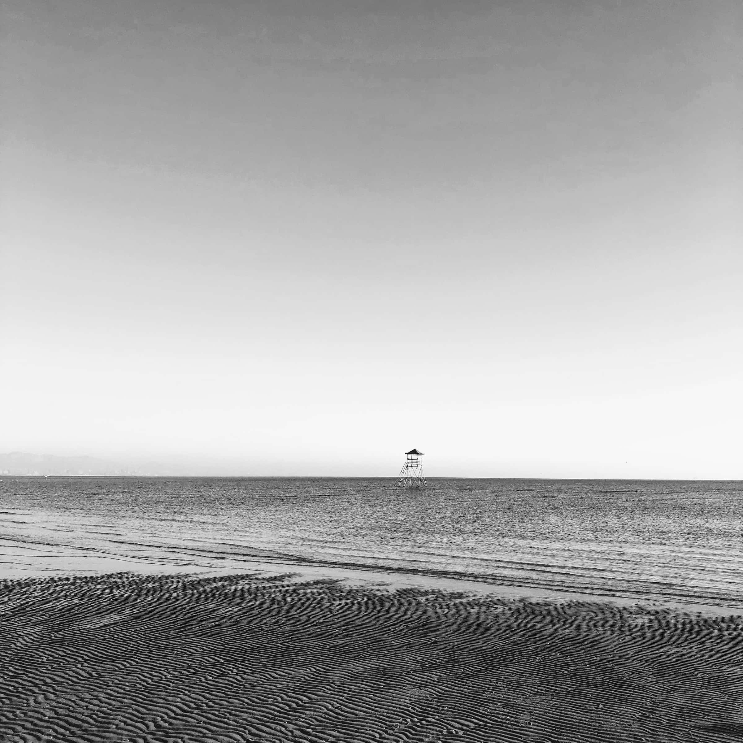 Black And White Beach 桌布 壁紙 — 一座孤獨的救生員塔矗立在廣闊、有紋理的沙灘上，背景是極簡的黑白天空。