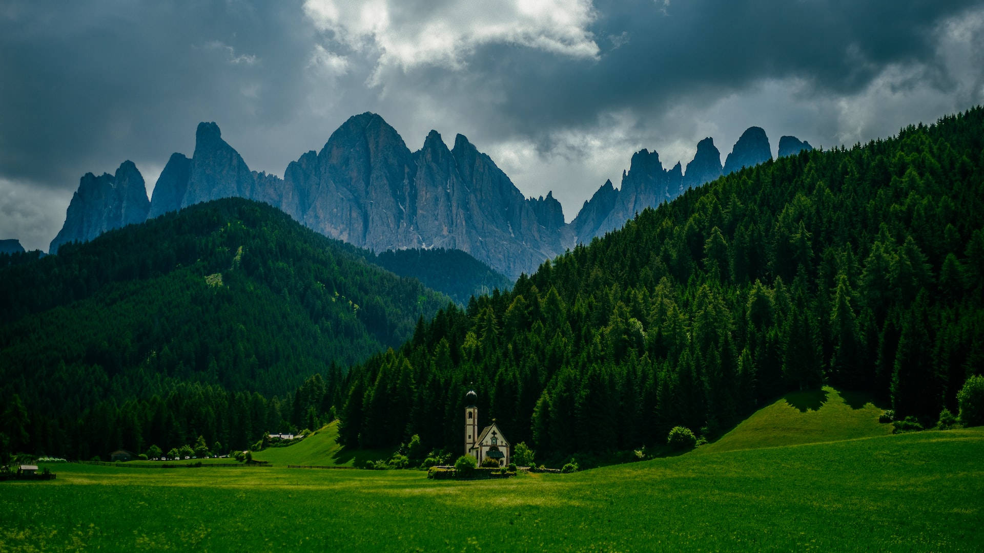 Mountain Landscape 桌布 壁紙 — 一座孤獨的教堂矗立在充滿活力的綠色草地上，下方是高聳的鋸齒狀山脈，上方是戲劇性的多雲天空。