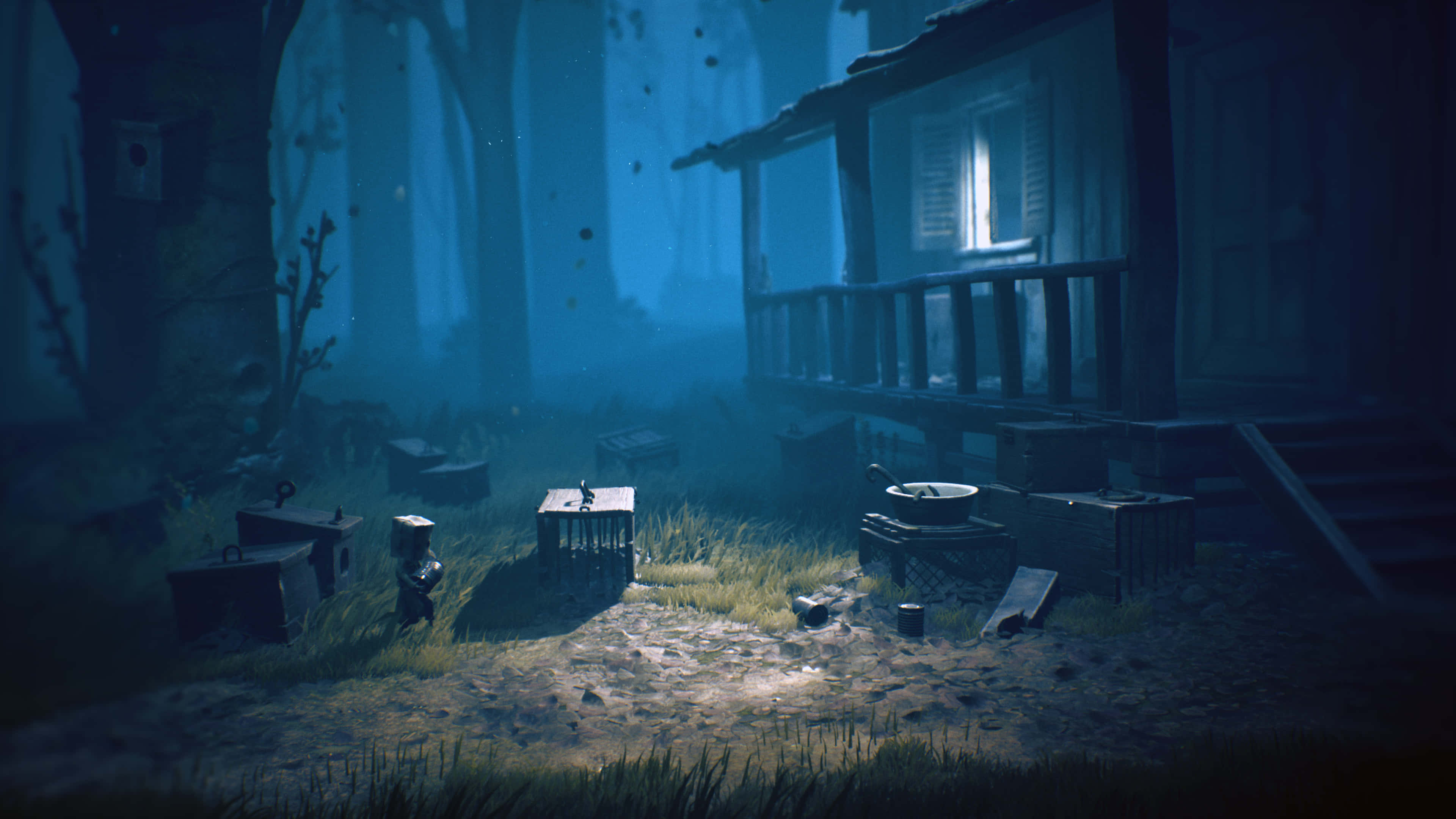 Little Nightmares 4K 桌布 壁紙 — 一座孤獨的木屋在迷霧森林中，從單一窗戶散發出溫暖的光芒，詭異的藍色霧氣在散落的板條箱和雜草叢生的草地上盤旋。
