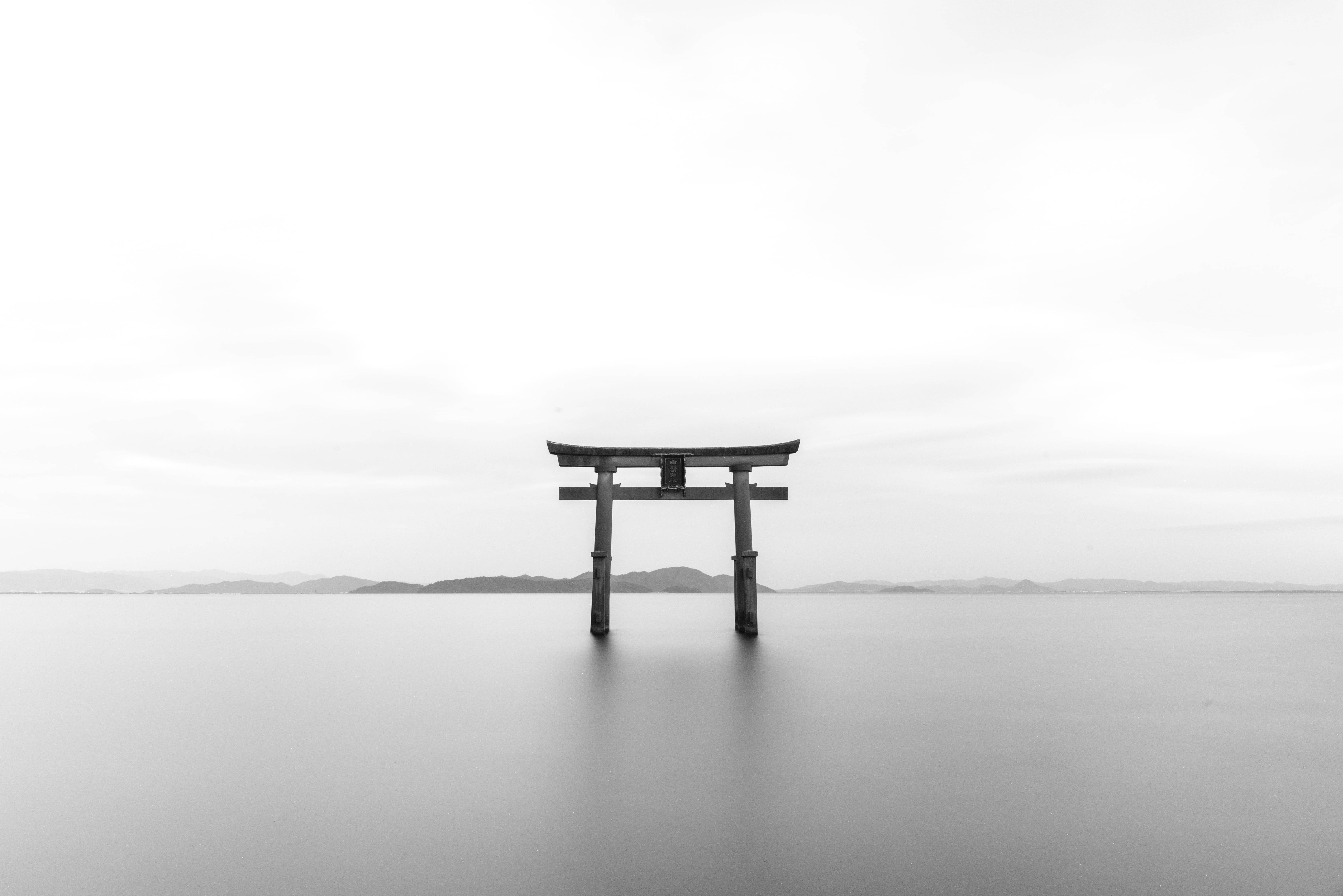 日本 桌布 壁紙 — 一座孤獨的木製鳥居矗立在平靜的水面上，天空多雲，遠方地平線上有山丘。