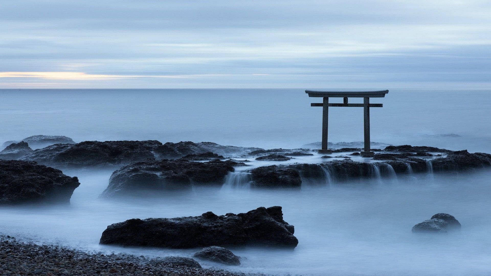 Torii 桌布 壁紙 — 一座孤獨的木製鳥居矗立在深色火山岩上，霧氣瀰漫的海水在柔和暮色天空下環繞流動。