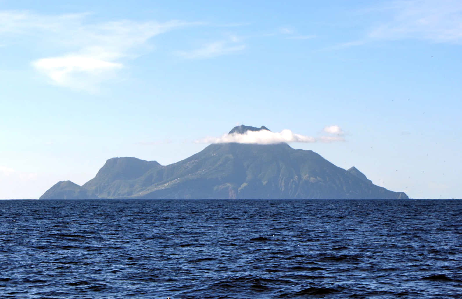 Scenery 桌布 壁紙 — 一座孤獨的森林火山島從深藍色的海洋中升起，天空淡藍，雲霧繚繞。