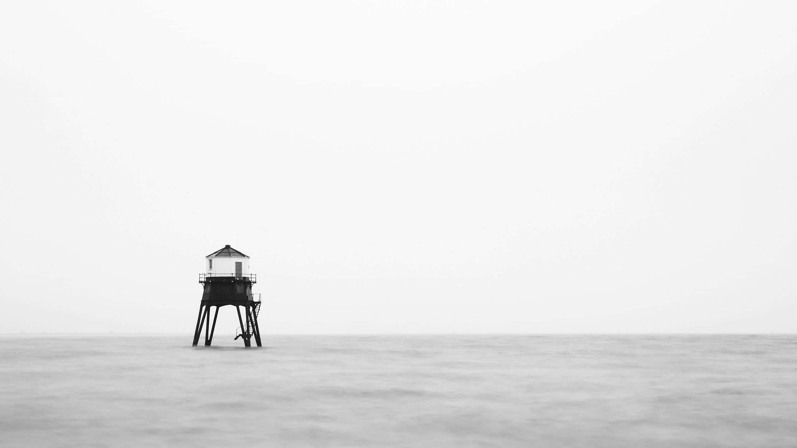 Black And White Ocean 桌布 壁紙 — 一座孤獨的燈塔矗立在廣闊、霧氣瀰漫的海洋中的支柱上，天空陰沉。