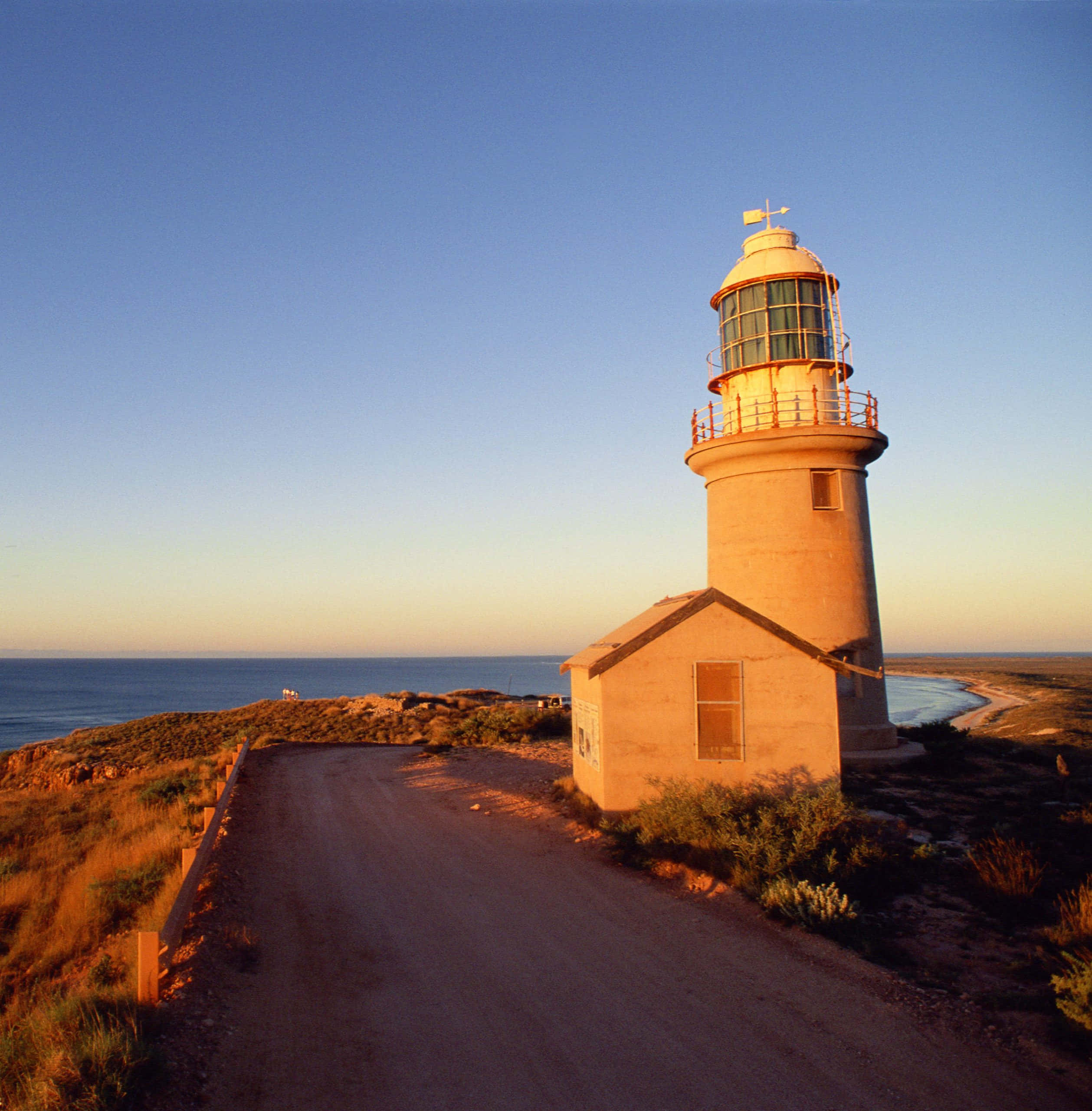 Lighthouse 桌布 壁紙 — 一座孤獨的燈塔矗立在海岸懸崖上，正值黃金時刻，溫暖的光線在蜿蜒的泥土路上投下長長的影子。