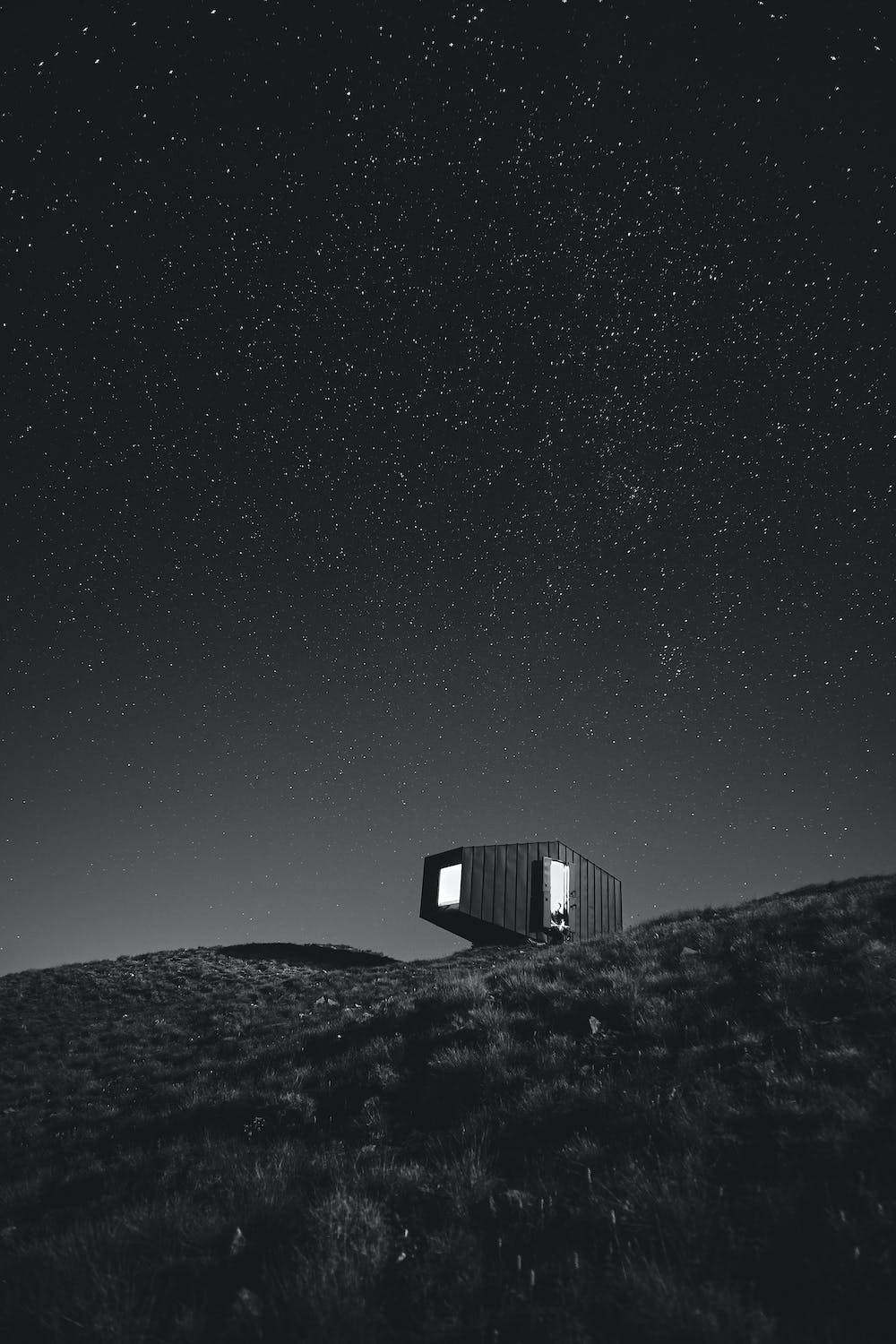 Stars 桌布 壁紙 — 一座孤獨的現代小屋坐落在長滿草的山丘上，下方是廣闊的星空夜空，窗戶閃閃發光。