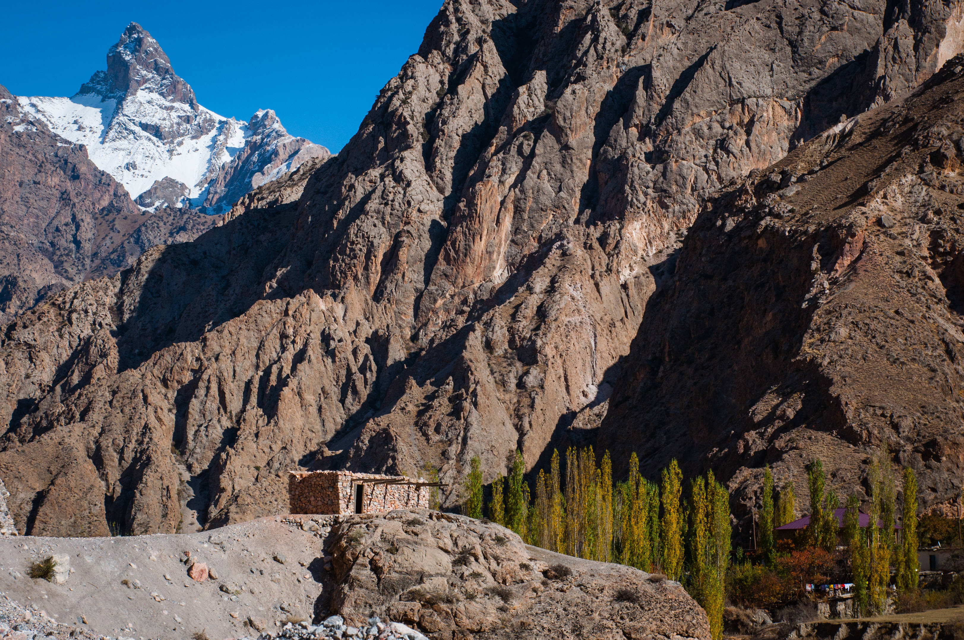Tajikistan 桌布 壁紙 — 一座孤獨的石屋棲息在岩石露頭上，下方是高聳的棕色懸崖，白雪覆蓋的山峰刺破清澈的藍天。
