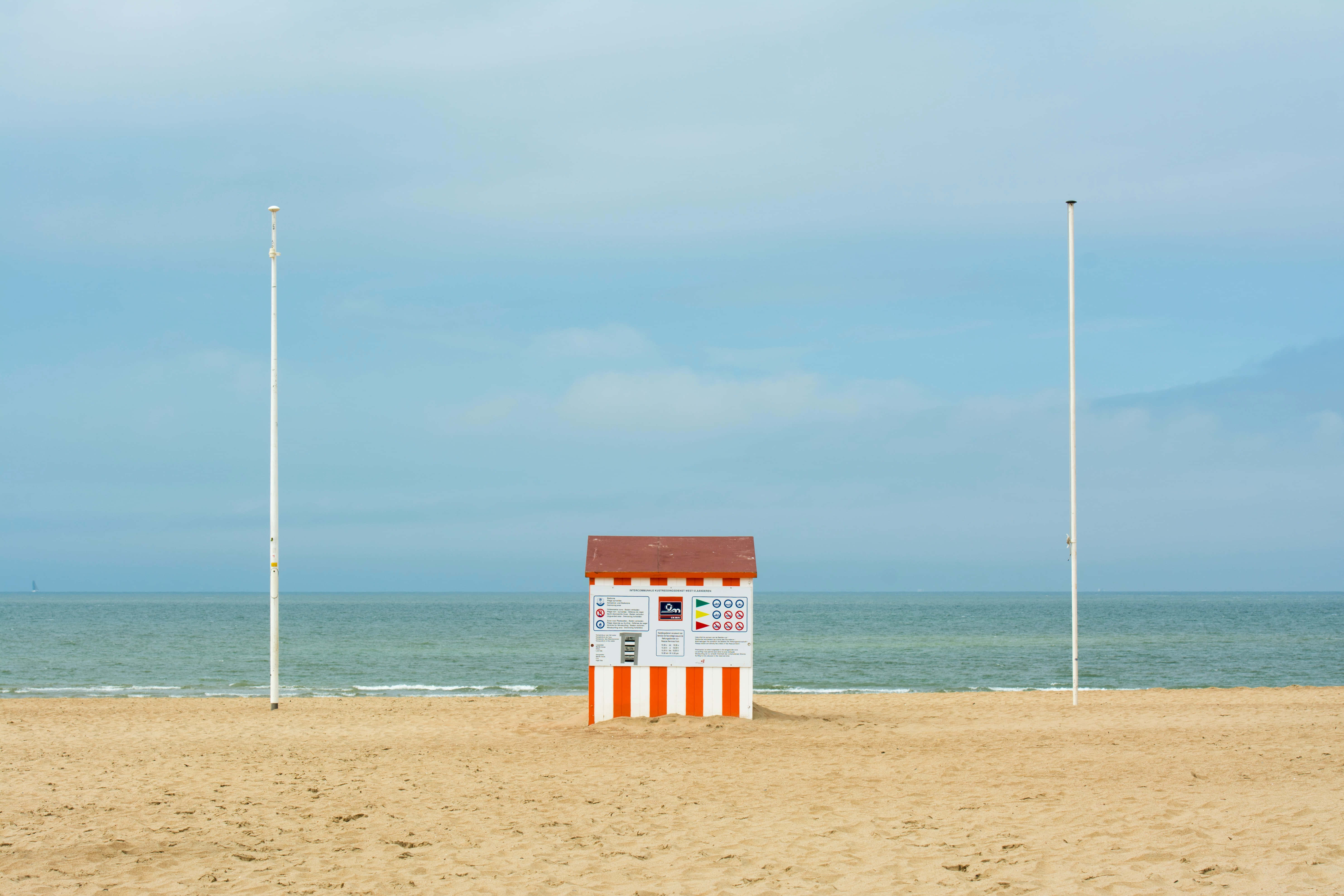 Aesthetic Beachy 桌布 壁紙 — 一座孤獨的紅白條紋救生員小屋矗立在寧靜的沙灘上，伴隨著平靜的海洋和多雲的天空。
