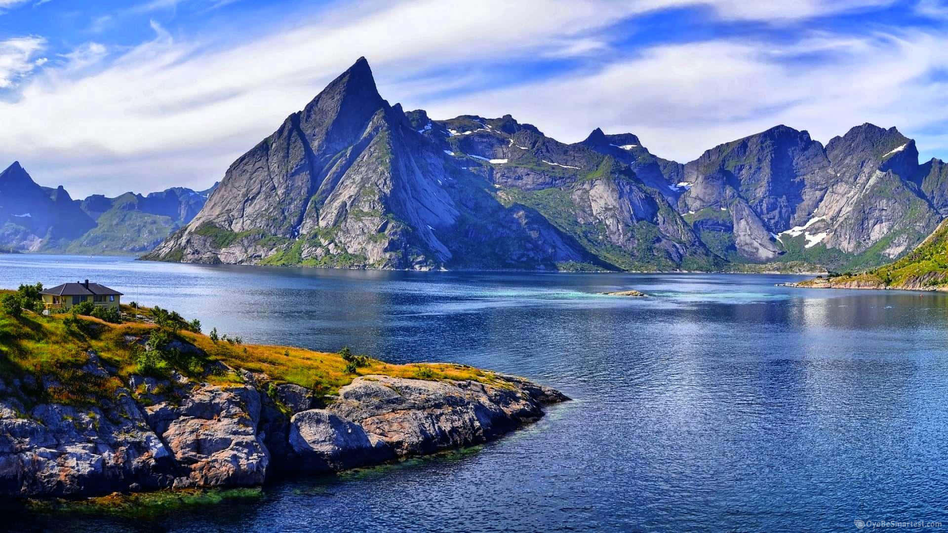 風景 桌布 壁紙 — 一座孤立的房子坐落在岩石海岸上，俯瞰著深藍色的峽灣，兩旁是高聳的鋸齒狀山脈，天空部分多雲。