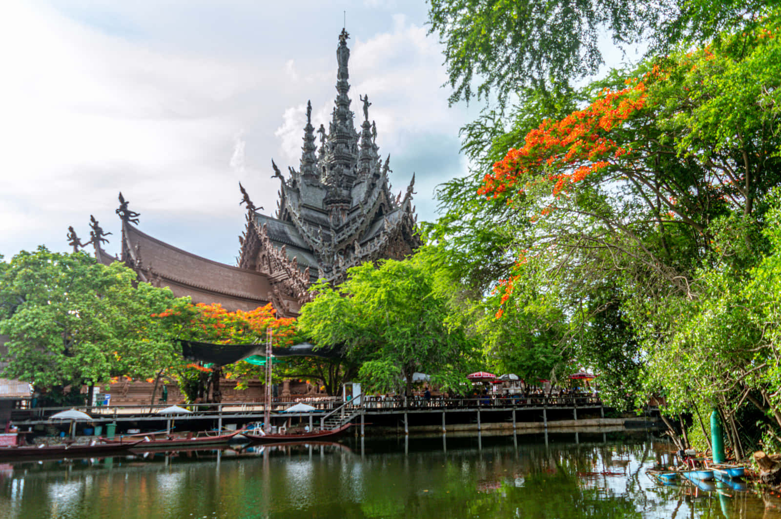 風景 桌布 壁紙 — 一座宏偉、雕刻精美的木造寺廟聳立在翠綠樹木和橙色花朵之後，倒映在平靜的水面上。
