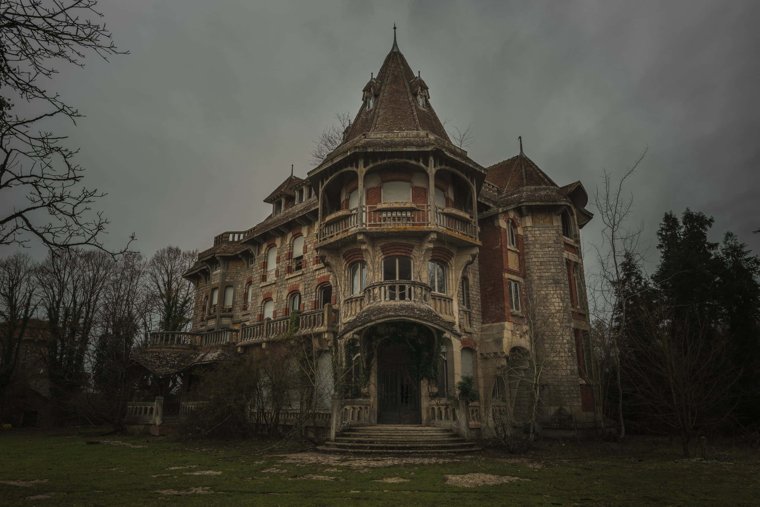 Haunted House 桌布 壁紙 — 一座宏偉但破敗的大宅，帶有華麗的陽台和石塔，矗立在陰沉多雲的天空下，周圍是光禿禿的樹木。