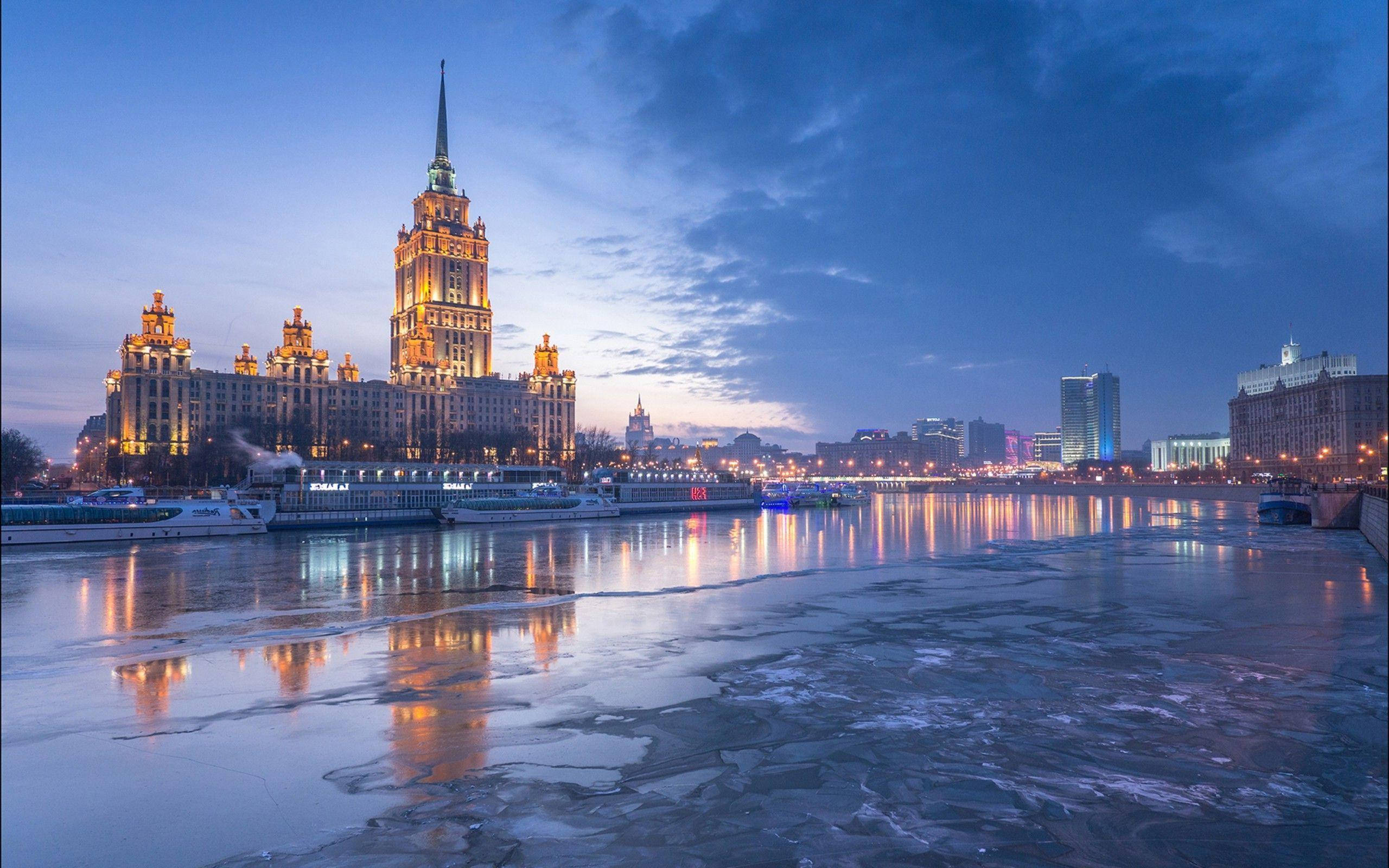 Moscow 桌布 壁紙 — 一座宏偉的、燈火輝煌的史達林式摩天大樓，在暮色蒼穹下倒映在一條部分結冰的河流上。