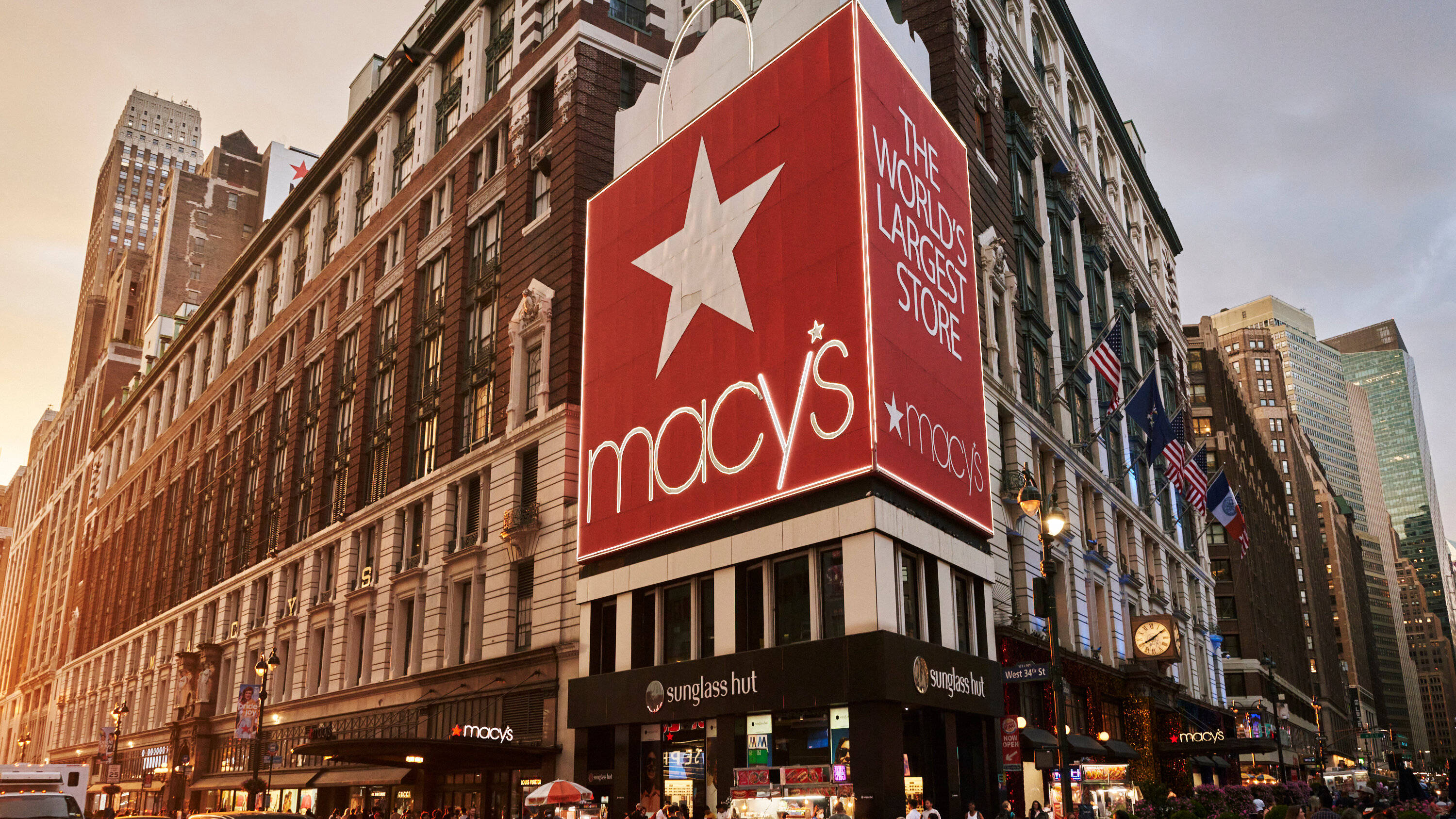 macys 桌布 壁紙 — 一座宏偉的Macy's百貨公司大樓，帶有巨大的紅色招牌和白色星星，在黃金時段主宰著繁忙的城市街道。