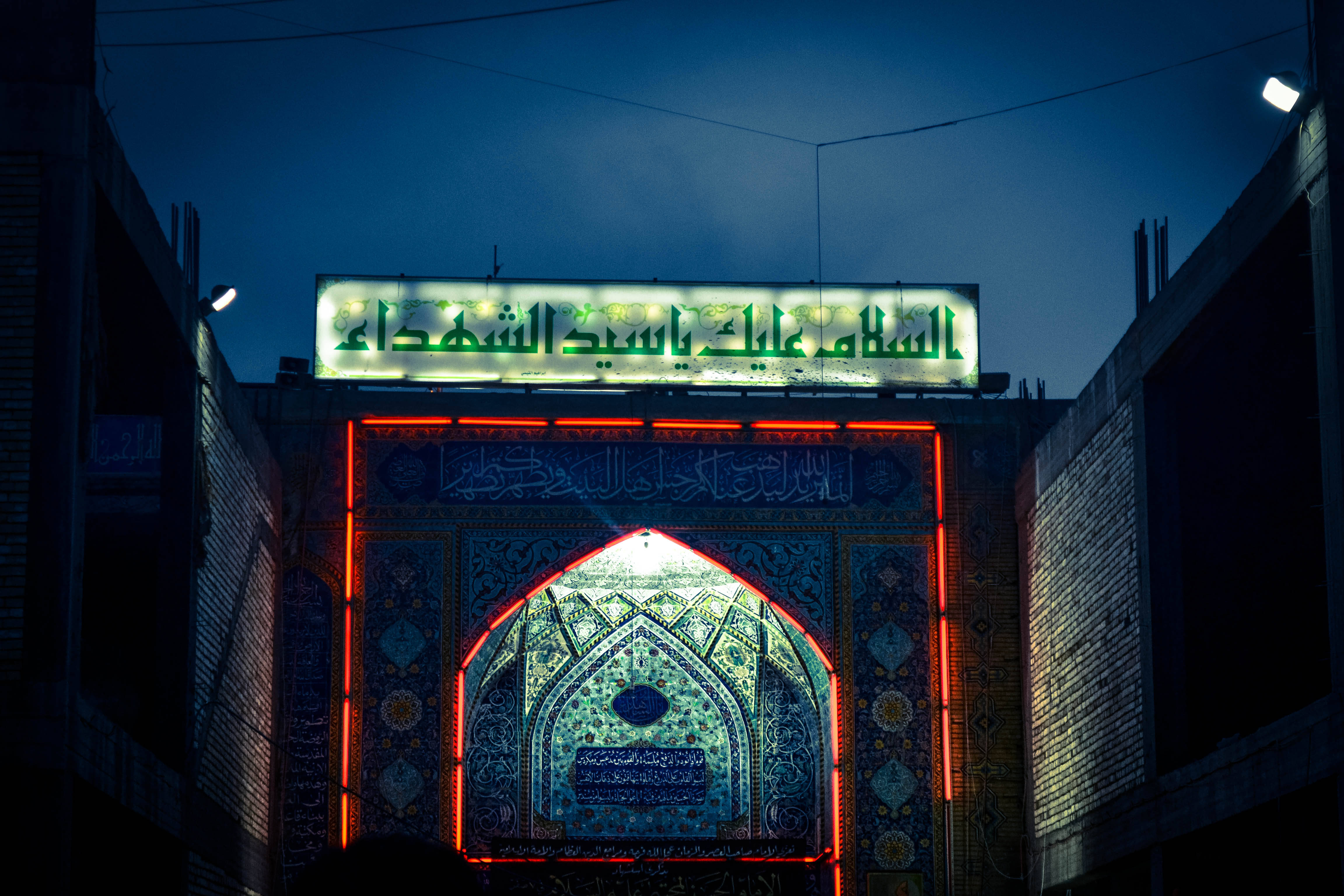 Karbala 桌布 壁紙 — 一座宏偉的伊斯蘭拱門在深沉的暮色天空下閃耀著紅色和藍色的燈光。