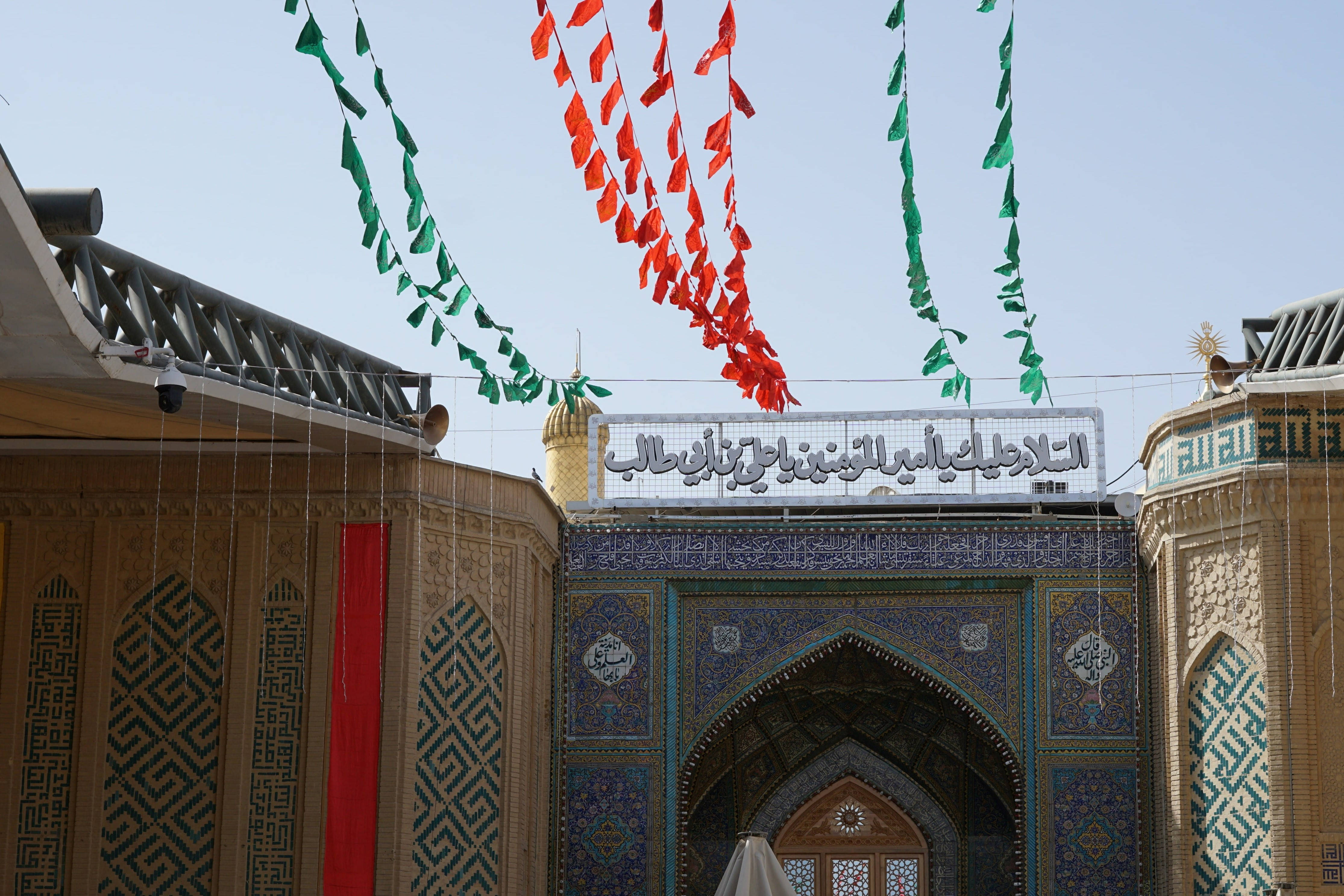 Iraq 桌布 壁紙 — 一座宏偉的伊斯蘭清真寺入口，飾有精緻的藍色瓷磚，紅綠旗幟在晴朗的天空下高高懸掛。