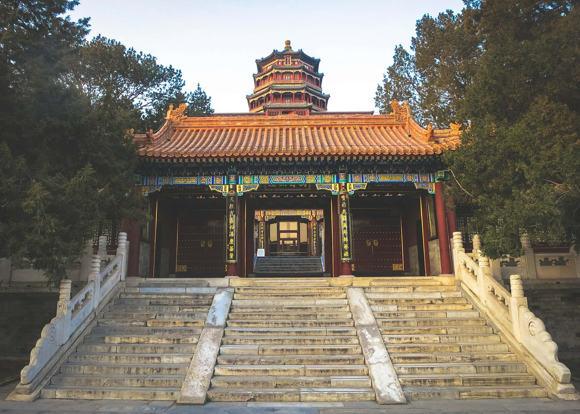 建築 桌布 壁紙 — 一座宏偉的傳統中國寺廟，擁有華麗的紅色和金色屋頂瓦片，矗立在寬闊的石階頂端，周圍環繞著高大的常青樹。