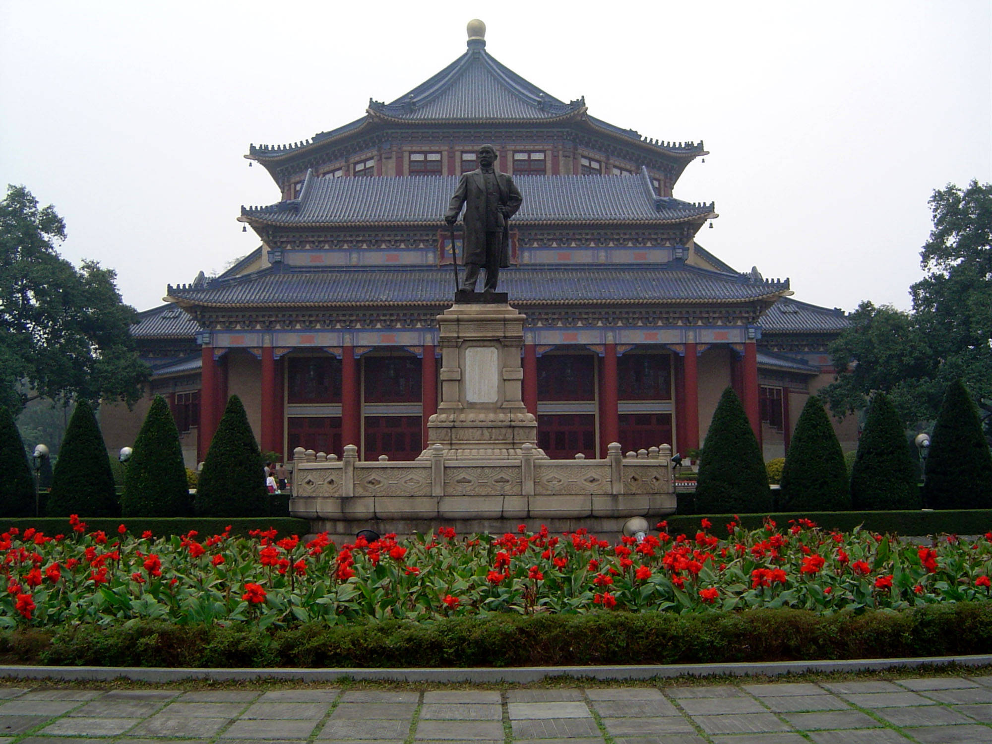 Guangzhou 桌布 壁紙 — 一座宏偉的傳統中式亭閣，帶有青銅雕像，矗立在色彩鮮豔的紅花壇後方，天空多雲。