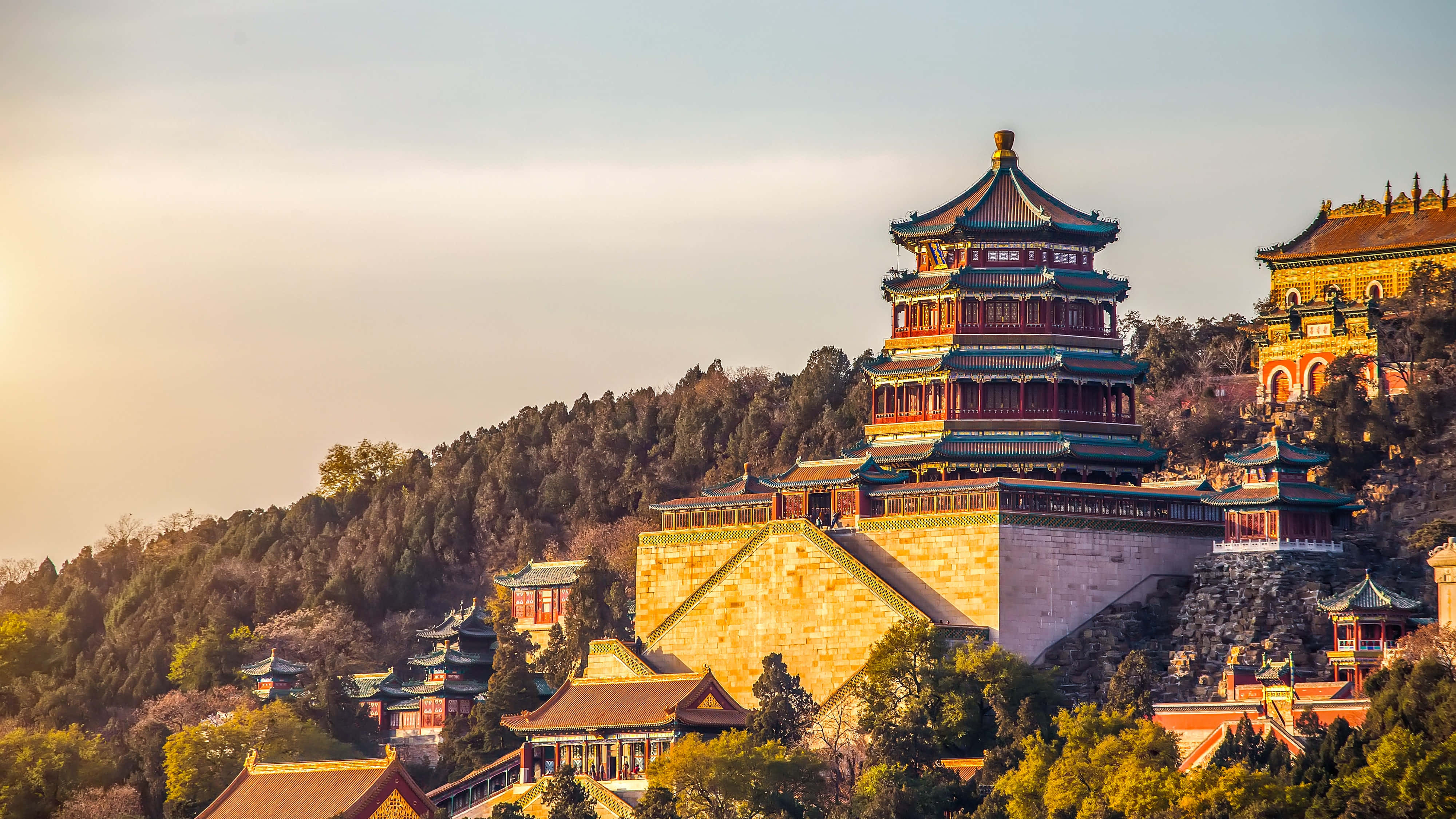 hill view 桌布 壁紙 — 一座宏偉的傳統中式寶塔，擁有分層屋頂和紅色點綴，雄偉地聳立在山坡上，周圍環繞著茂密的綠樹，在柔和的金色日落天空下。